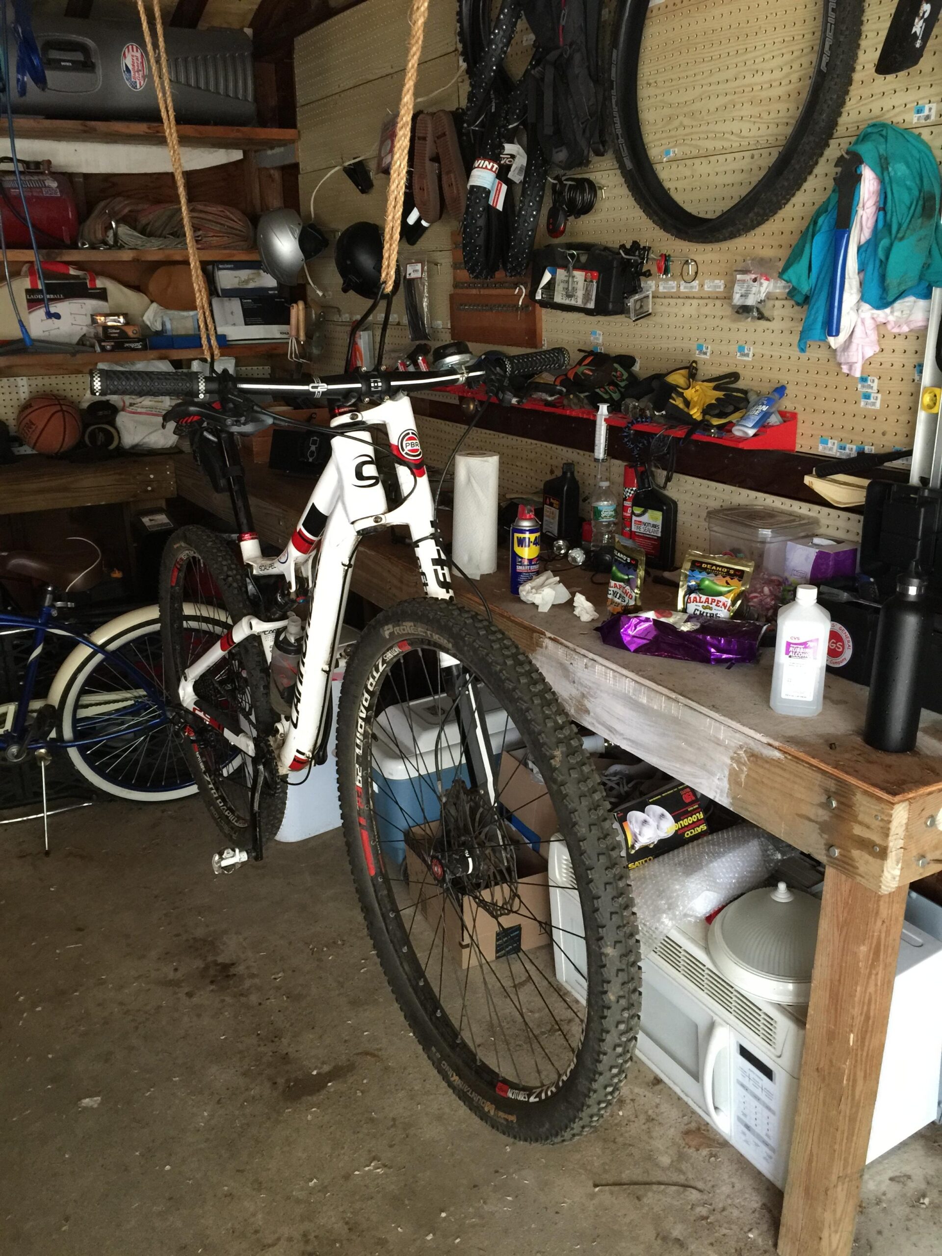 Cannondale Scalpel 29er 3: A mountain bike hanging in a cluttered garage, with various tools, equipment, and sports items scattered around. The bike is suspended from the ceiling by ropes, and the walls are lined with helmets, tires, and clothing. A workbench is visible in the foreground, filled with miscellaneous items such as cleaning supplies and packaging materials.