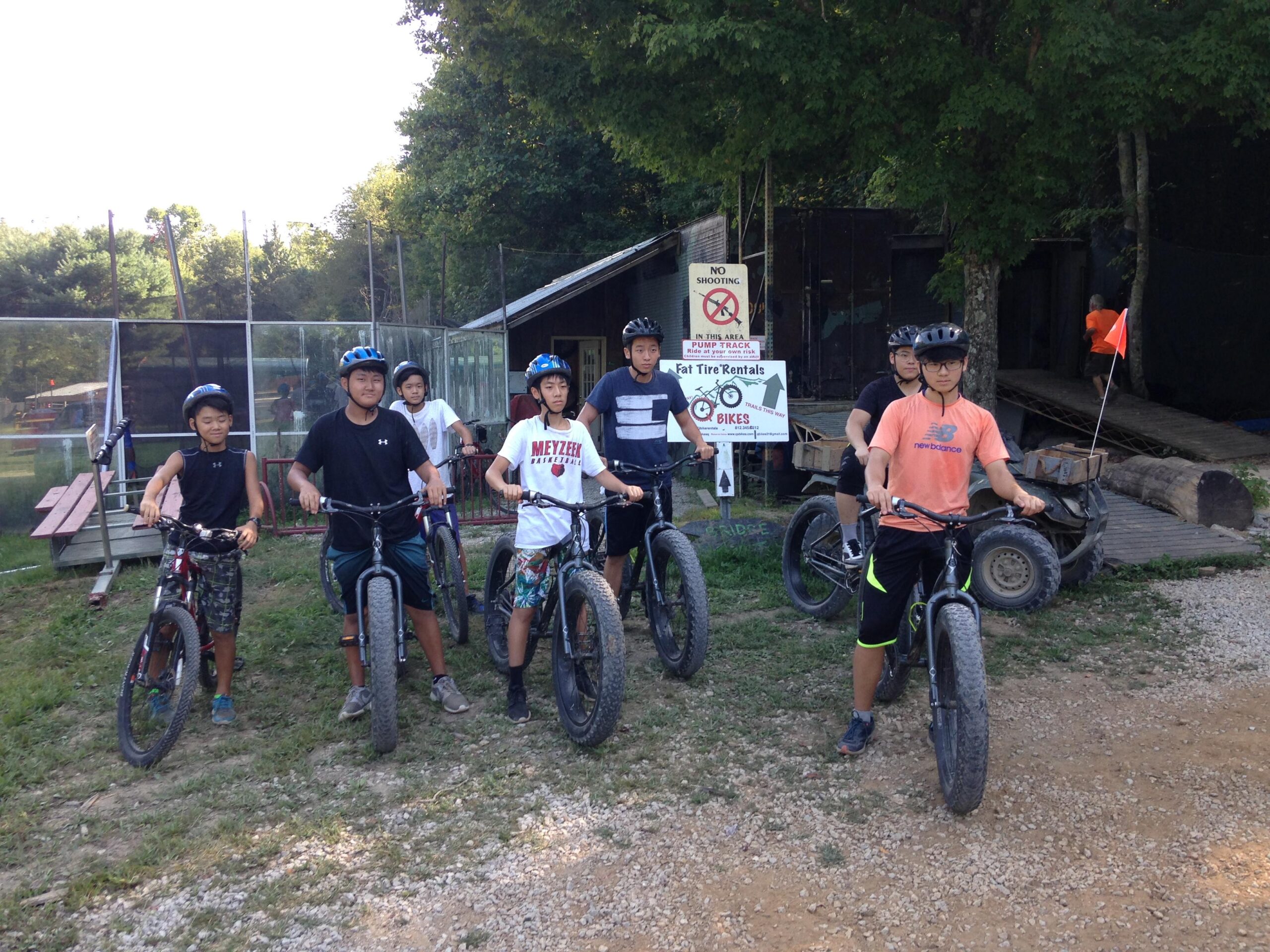 A group of six children in casual clothing and wearing helmets, standing on fat tire bicycles in an outdoor setting. Behind them, there is a sign indicating "No Shooting" and "Fat Tire Rentals." The background features trees and a wooden structure, suggesting they are at a recreational area or bike park.