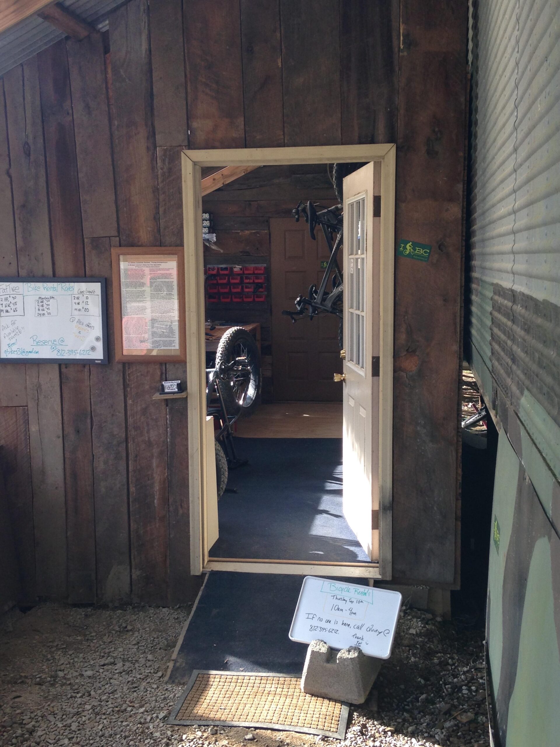 A rustic wooden building with an open door leading inside, showcasing a welcoming ambiance. There is a sign outside on the ground that provides information about bicycle rentals, including hours of operation. Inside the entrance, bicycles are visible hanging on the wall, and a bulletin board displays additional information. The floor is carpeted, and the space appears well-lit and organized.