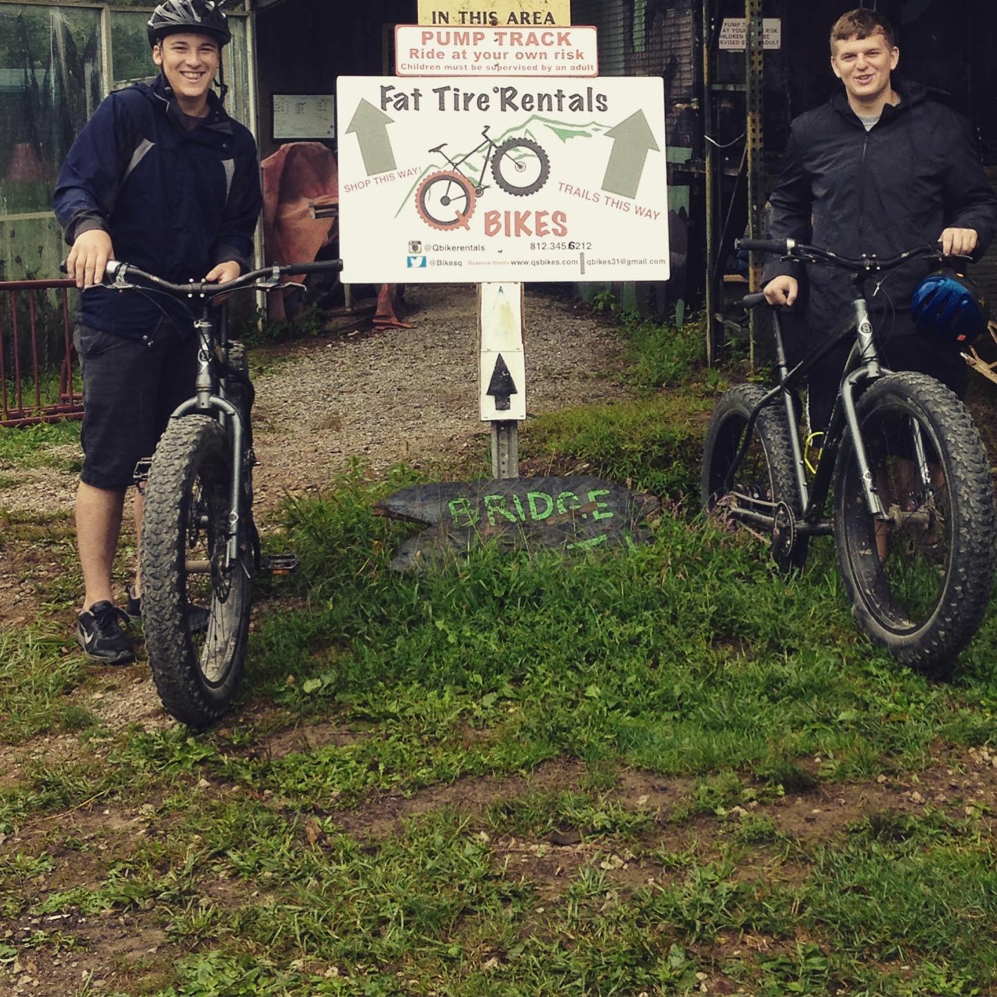 Two individuals are standing beside a sign for "Fat Tire Rentals" at a bike rental station. They are each holding a fat tire bike and are wearing casual outdoor clothing. The sign indicates directions for "Shop This Way" and "Trails This Way," and it also features a warning about riding at one's own risk. The background includes greenery and a gravel path, setting a recreational outdoor vibe.