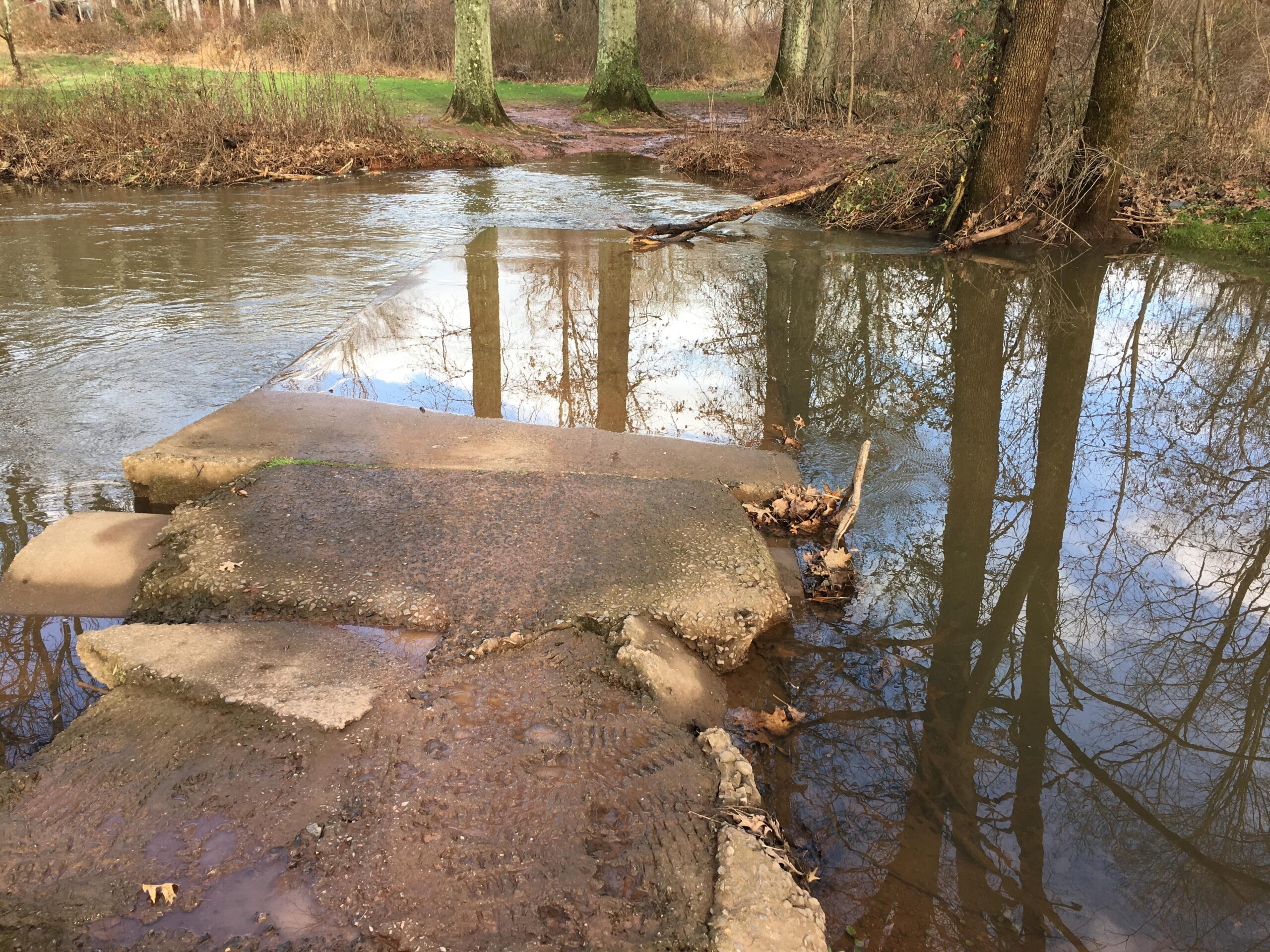 A rocky pathway leading to a shallow, flowing stream, surrounded by trees and reflecting the sky. The water appears calm, with some brown leaves scattered near the edge. Six Mile Run mountain bike trail.