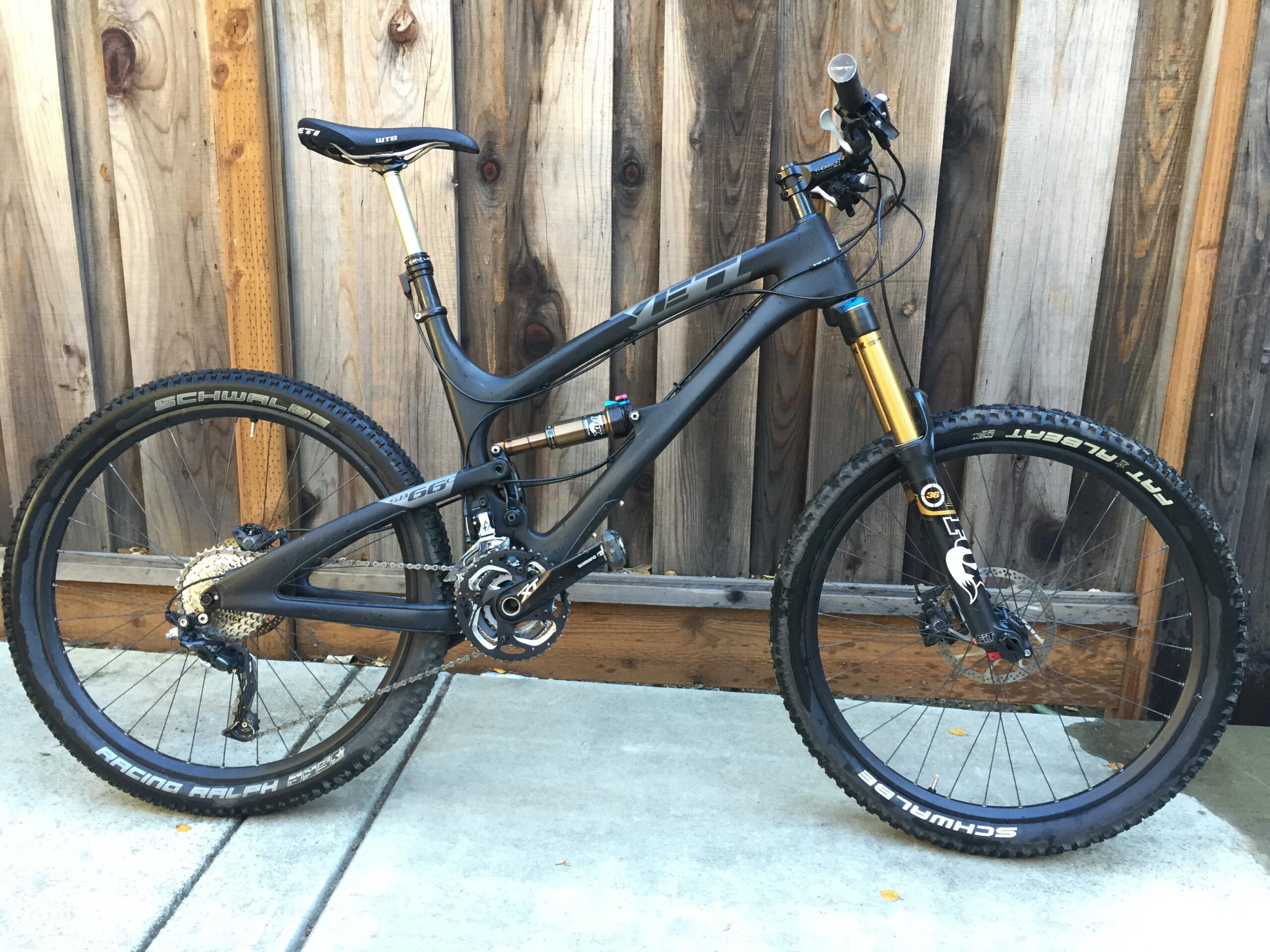 Yeti SB66: Image of a mountain bike with a black frame and gold suspension fork, parked against a wooden fence. The bike features wide tires with visible tread, a dropper seatpost, and a chain drivetrain system. The background consists of rustic wooden panels.