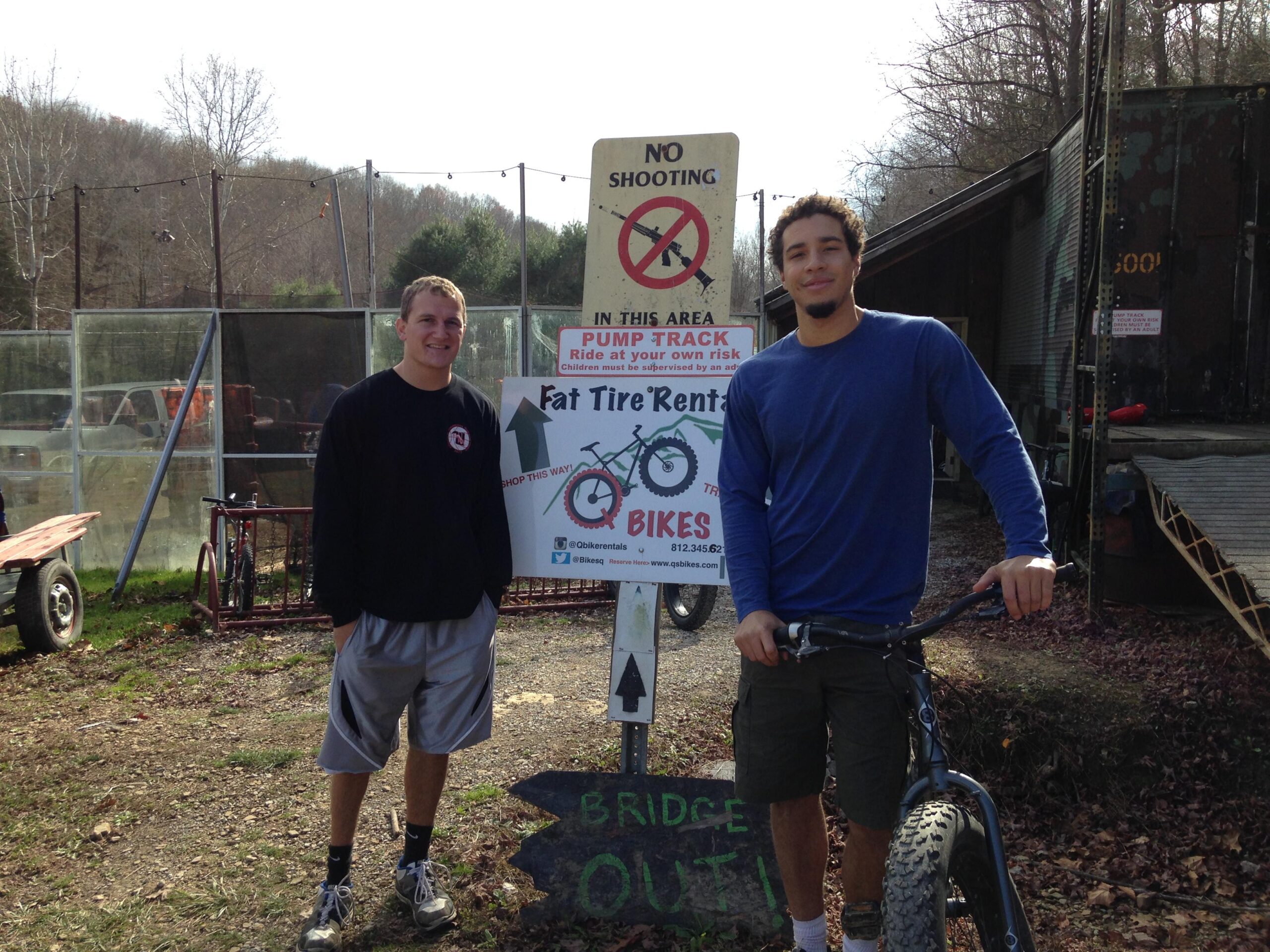 Two young men stand in front of a sign that reads "No Shooting in this Area" and provides information about bike rentals, specifically for fat tire bikes. One man is wearing a black long-sleeve shirt and gray shorts, while the other is in a blue long-sleeve shirt and shorts. They are positioned next to a bike, with trees and a structure in the background. The setting appears to be an outdoor recreational area.