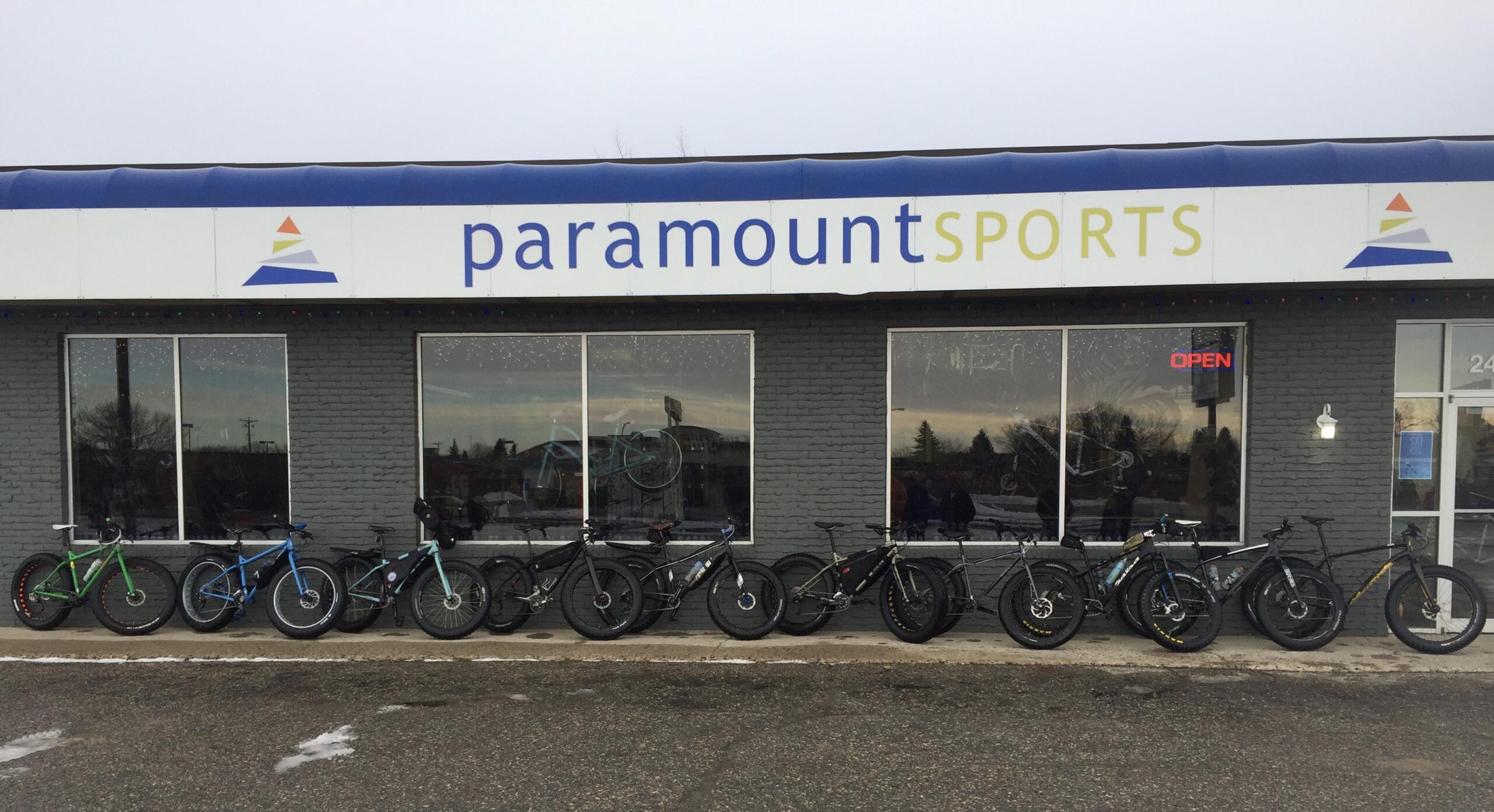 Alt text: A row of bicycles lined up outside a store with the sign "paramount SPORTS." The store features large windows and a visible "OPEN" sign. The bicycles vary in color, including green, blue, and black, positioned along the sidewalk in front of the store.