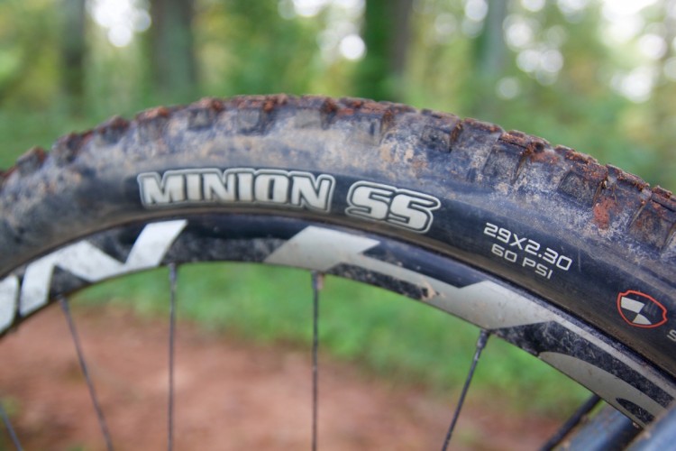 Close-up view of a dirty mountain bike tire labeled 'MINION SS' with specifications showing '29x2.30' and '60 PSI,' set against a blurred green forest background.
