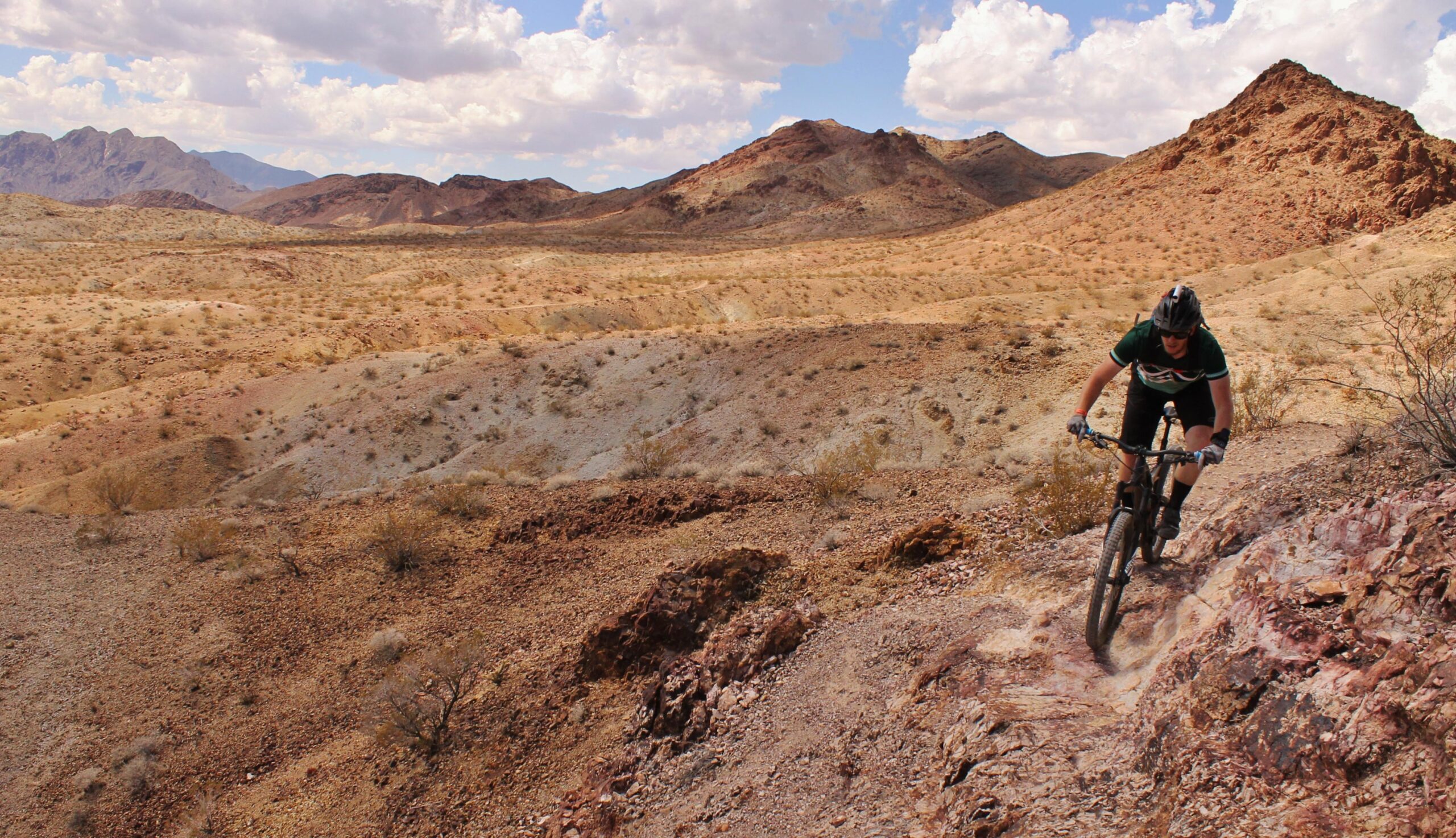 A mountain biker navigating a rocky trail in a desert landscape with rolling hills and distant mountains under a partly cloudy sky. Bootleg Canyon mountain bike trail.