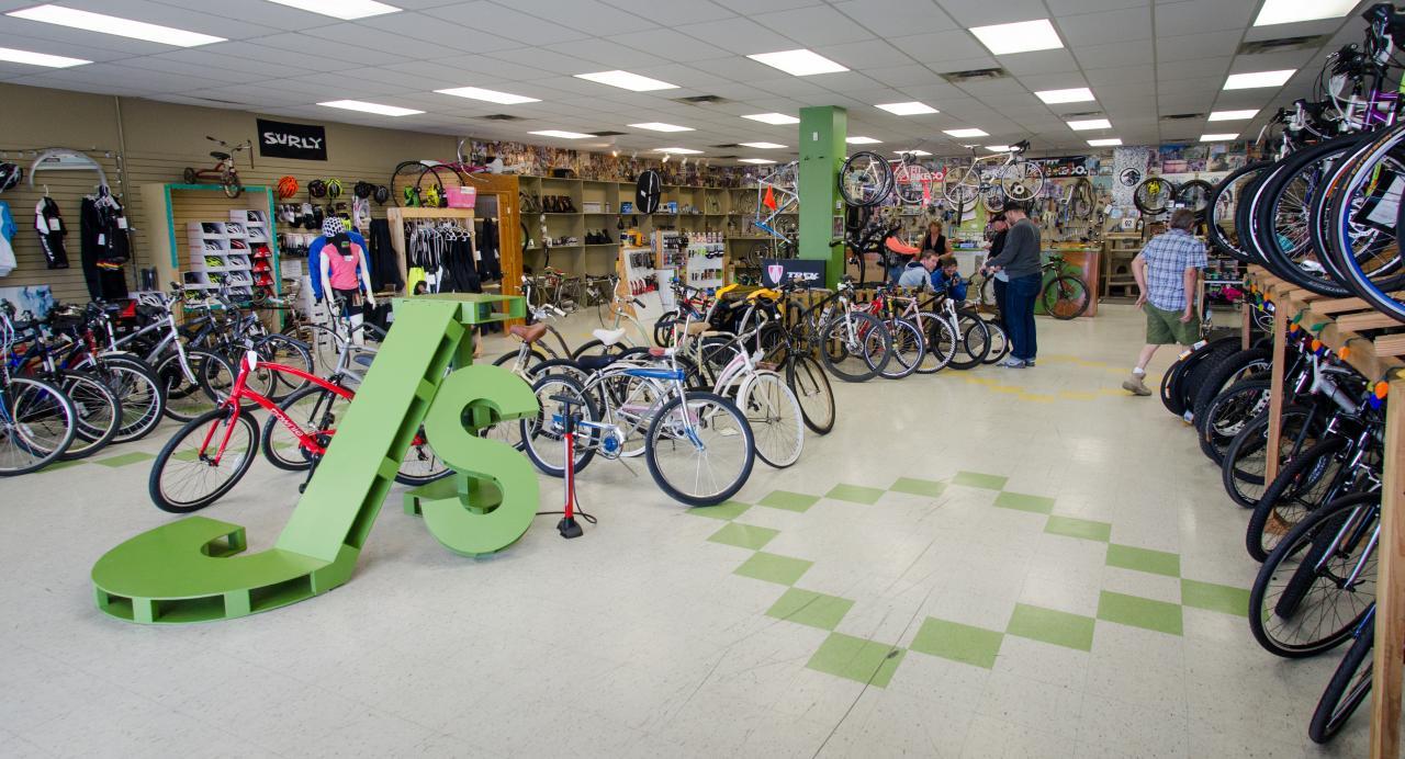 A lively bike shop interior featuring various bicycles for sale, a large green "J's" sign prominently displayed in the foreground, and customers browsing among displays of bikes and cycling gear. The shop is well-lit with organized shelves showcasing accessories and apparel, while families and individuals interact and shop throughout the space.