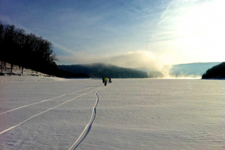 Two people walking on a snowy lake with bicycle tracks in the fresh snow, surrounded by misty mountains and a clear blue sky.
