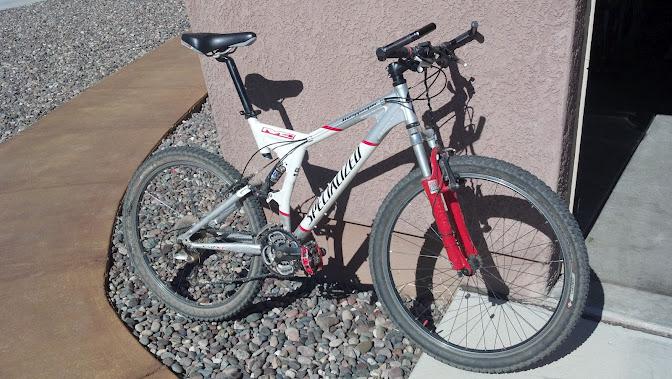 Specialized Stumpjumper FSR: A white and red mountain bike parked on a concrete surface, with a rocky ground and a wall in the background. The bike features thick tires and front suspension, indicating it is designed for off-road riding.