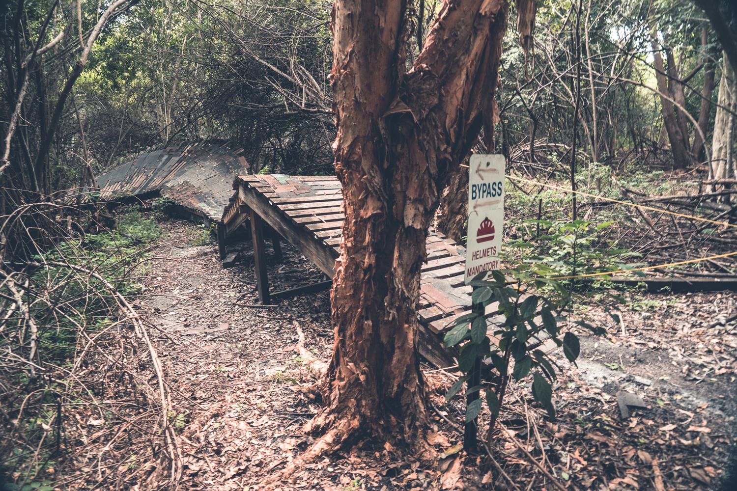 An overgrown path in a dense forest features a deteriorating wooden structure, partially collapsed to one side. A sign indicating "BYPASS" and "HELMETS MANDATORY" is visible next to a tree, surrounded by thick underbrush and fallen leaves. Markham Park mountain bike trail.
