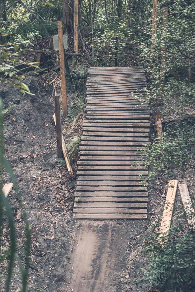 A wooden bike ramp winds through a forested area, surrounded by greenery and dirt paths. The ramp is built from planks and is elevated, leading into a path that creates a bridge-like structure. The scene is shaded with trees and foliage, showcasing a rustic and natural setting. Markham Park mountain bike trail.