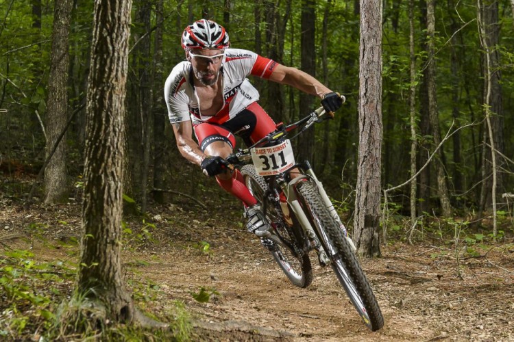 A male mountain biker in a racing outfit rides aggressively on a dirt trail surrounded by trees. He is leaning into a turn, showcasing his skill as he navigates the terrain. The bike features a visible race number 311. The background is a lush green forest, indicating a competitive mountain biking event.