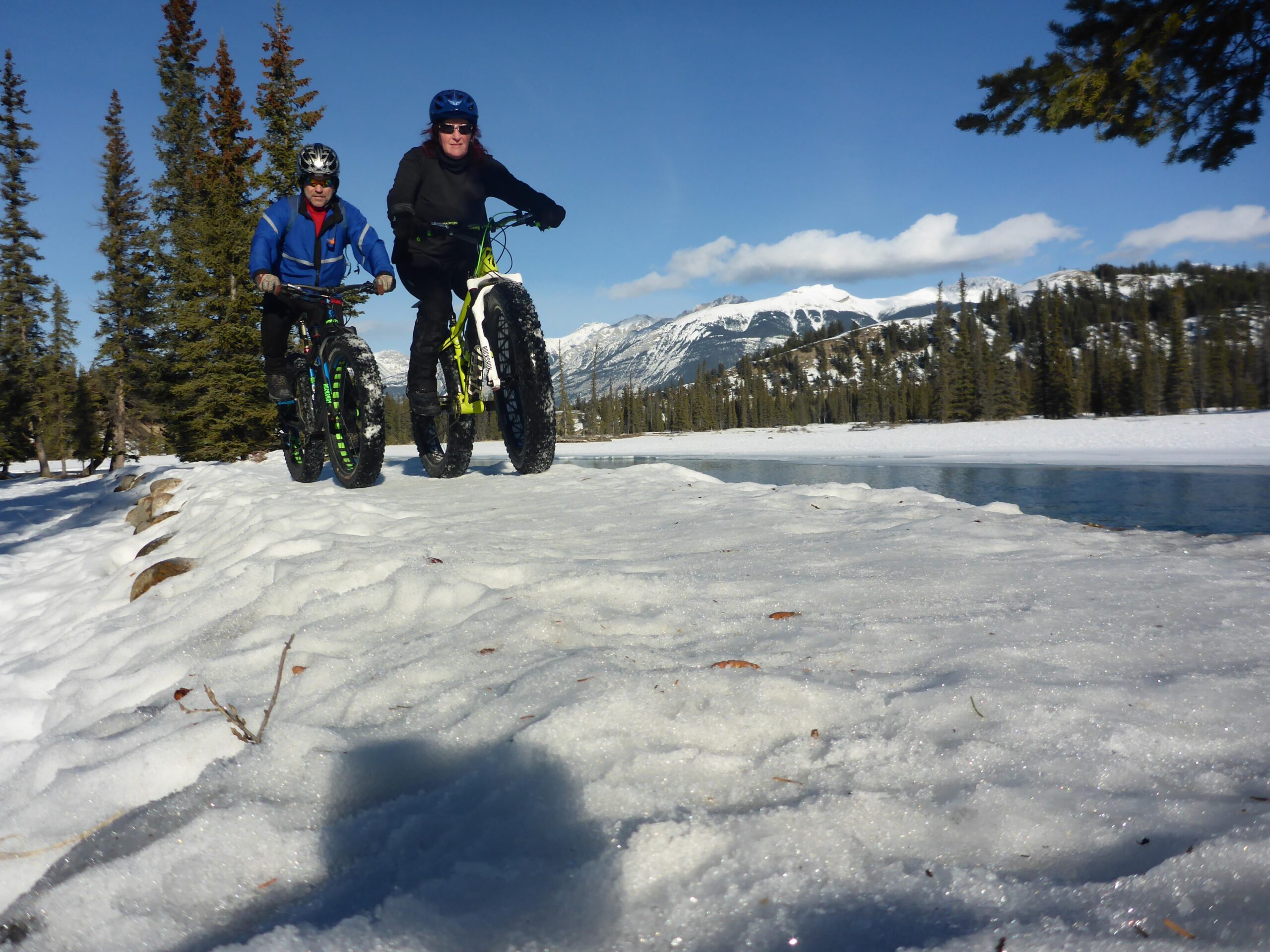 Two people riding fat bikes along a snowy path beside a frozen lake, surrounded by tall evergreen trees and mountains in the background under a clear blue sky. 12 to 5 to 5 / Valley of the Five Lakes and Wabasso Lake mountain bike trail.