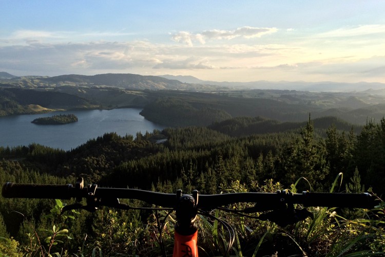 A mountain bike is positioned in the foreground, with its handlebars visible, overlooking a scenic vista of a tranquil lake surrounded by lush green hills and forests. The sky is clear, with soft clouds, and the sun casts a warm glow over the landscape, indicating either early morning or late afternoon light.