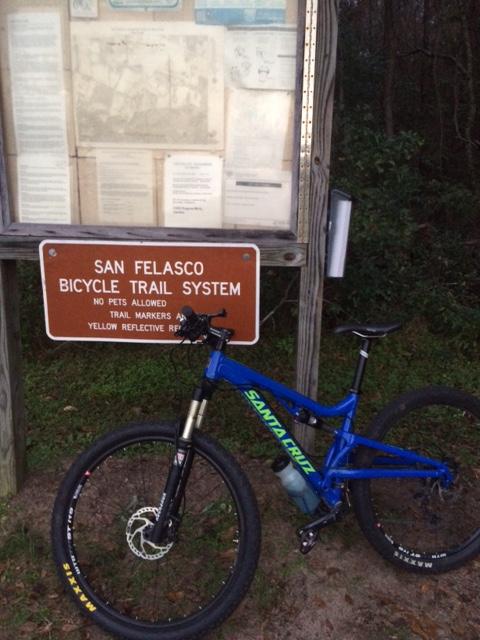 A blue mountain bike rests next to a sign for the San Felasco Bicycle Trail System. The sign features information about the trail, including rules such as "No pets allowed" and details about trail markers. Behind the bike, there are informational posters and maps related to the trail system. The setting is wooded, indicating a natural outdoor environment. San Felasco Hammock Preserve mountain bike trail.