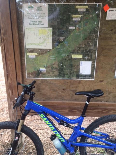 A mountain bike leaning against a wooden sign that features a detailed map of the Santos Bike Trailhead East, part of the Marjorie Harris Carr Cross Florida Greenway Recreation and Trails. The map highlights various trails and points of interest in the area. Santos mountain bike trail.