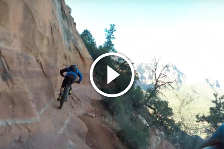 A mountain biker navigating a rocky trail on a steep cliffside, surrounded by lush greenery and distant mountains in the background. The focus is on the rider in a blue jacket as they maneuver along the rugged terrain.