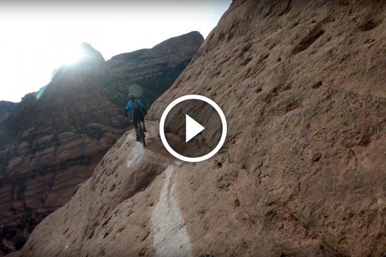 A mountain biker riding along a narrow trail on a rocky cliffside with mountains and a bright sun in the background. A play button overlay indicates this is a video.