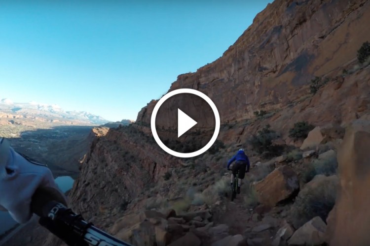 A mountain biker navigating a rocky trail along a canyon, with a scenic view of mountains and a river below, against a clear blue sky.