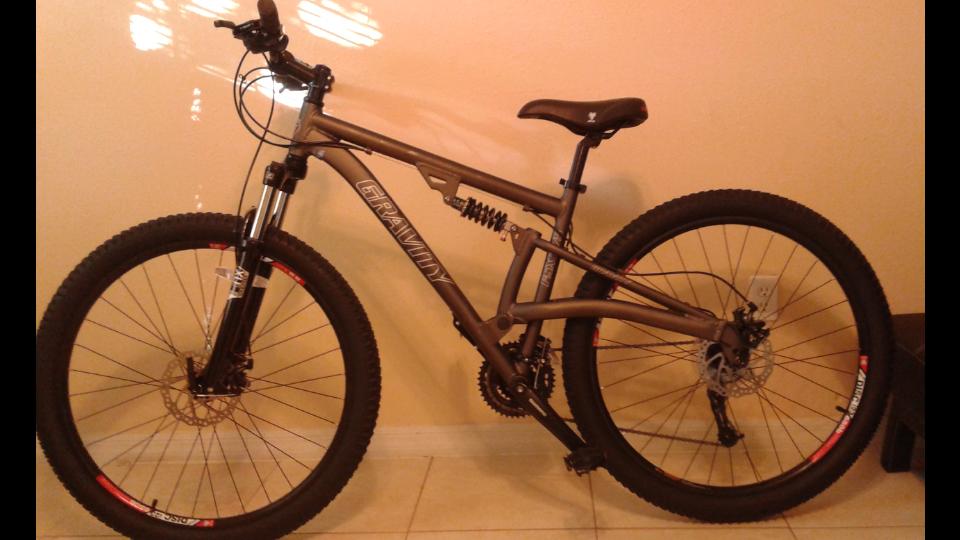 Gravity FSX2750 DS: A gray mountain bike with thick tires and a sturdy frame, featuring front suspension and a prominent brand logo. The bike is positioned against a beige wall, showcasing its design and structure.