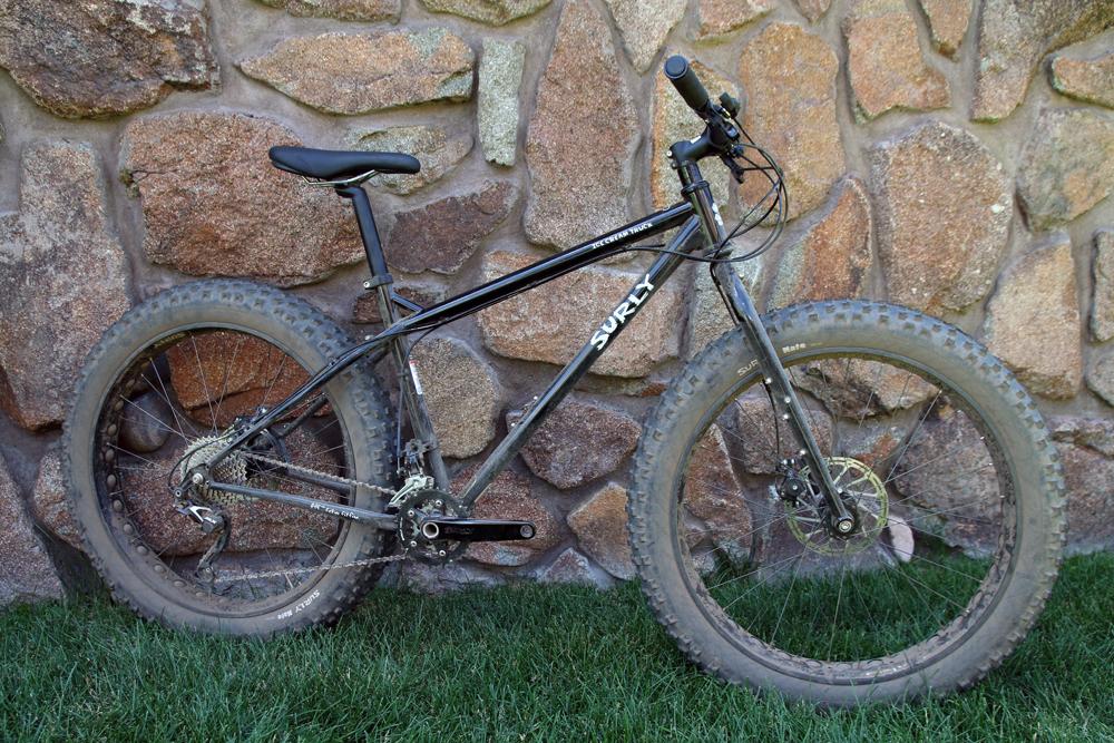 Surly Ice Cream Truck Ops: A black Surly fat bike is leaning against a textured stone wall, showcasing its wide tires and rugged frame. The bike is partially covered in dirt, indicating it has been used for off-road riding. The scene is set on a grassy area, contrasting with the stone background.