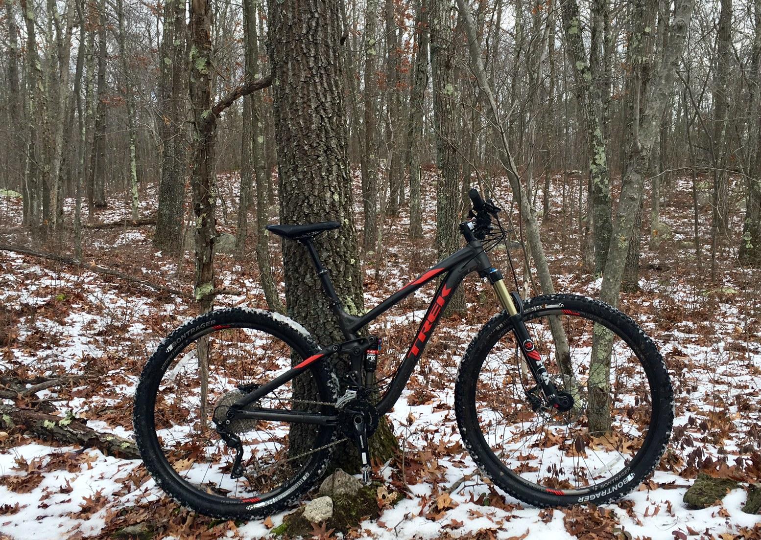 Trek Fuel EX 8: A black and red mountain bike leaning against a tree in a snowy forest. The ground is covered with fallen leaves and a light layer of snow, with trees in the background creating a serene, wooded setting.