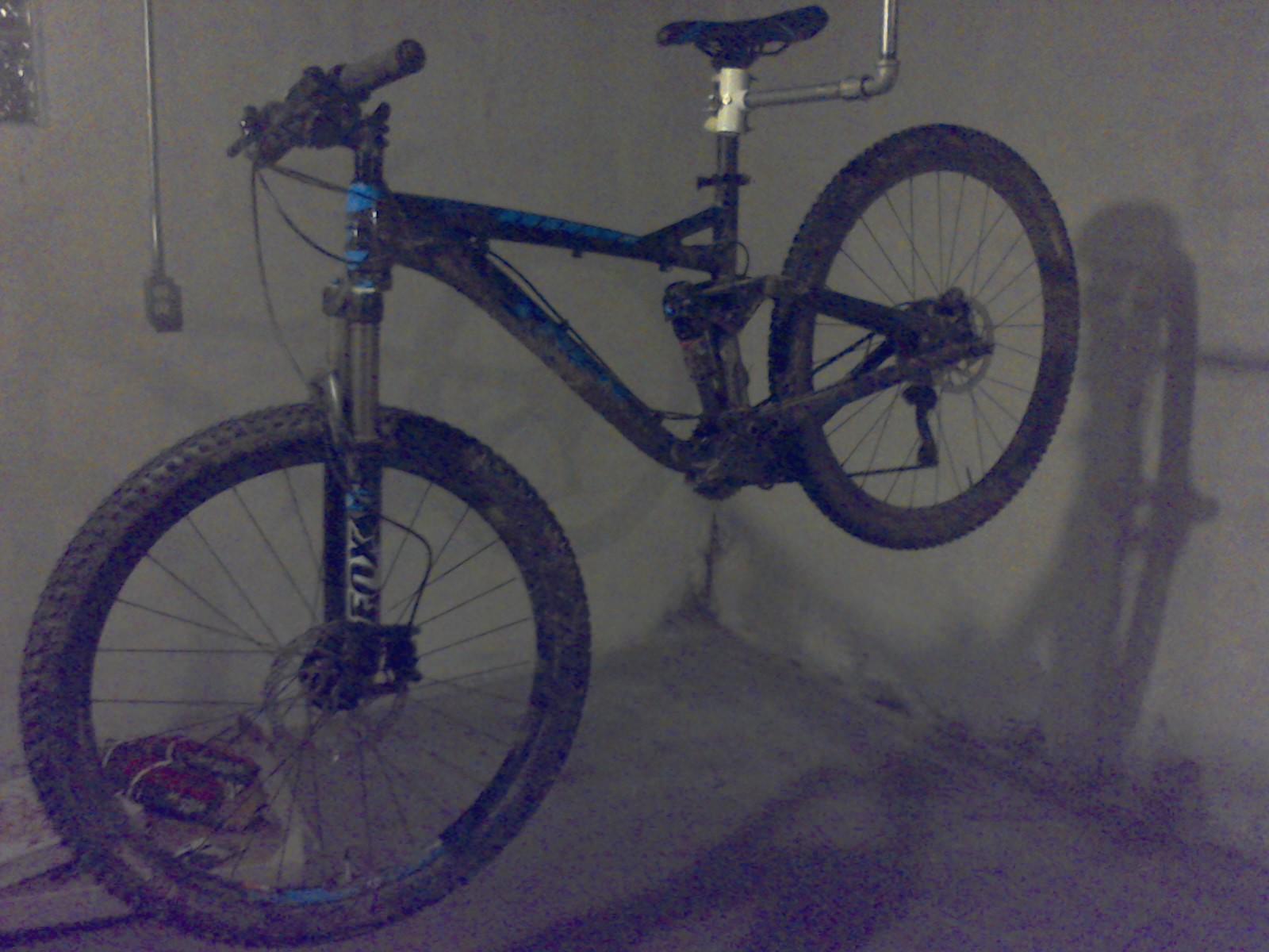Trek Fuel EX 7: A blue and black mountain bike hanging in a dimly lit garage with a concrete floor. The bike appears to be well-used, with dirt on the tires and frame.