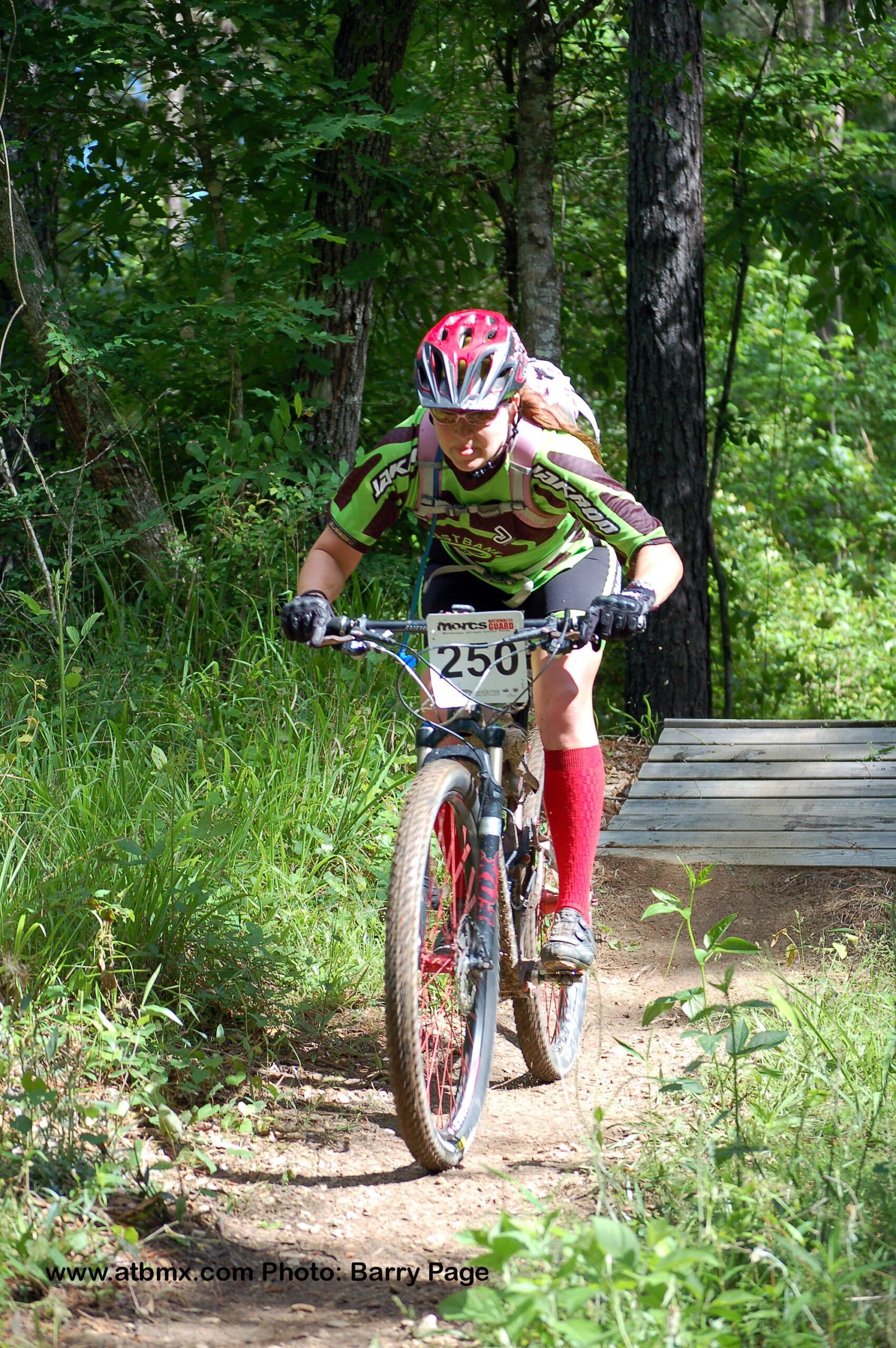 Niner RIP 9: A mountain biker wearing a vibrant green and brown jersey and red socks navigates a dirt trail surrounded by trees. The cyclist is focused and leaning forward on the bike, approaching a wooden ramp. The scene captures the energy and excitement of outdoor biking.