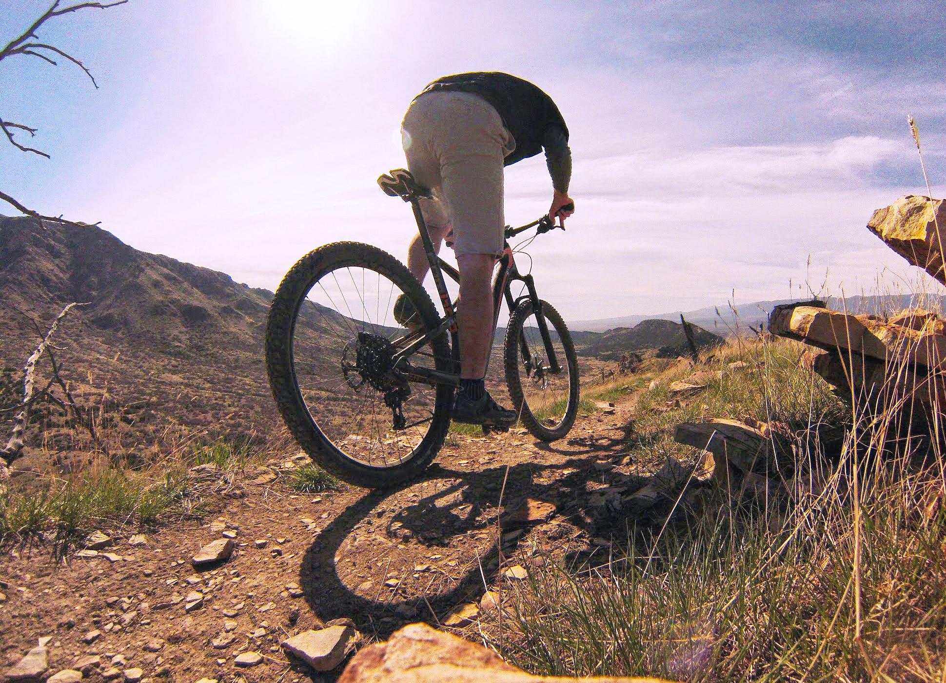 A person riding a mountain bike on a dirt trail surrounded by rocky terrain and hills, with sunlight shining in the background. The rider is focused on navigating the path, with grass and rocks visible along the trail. Oil Well Flats mountain bike trail.