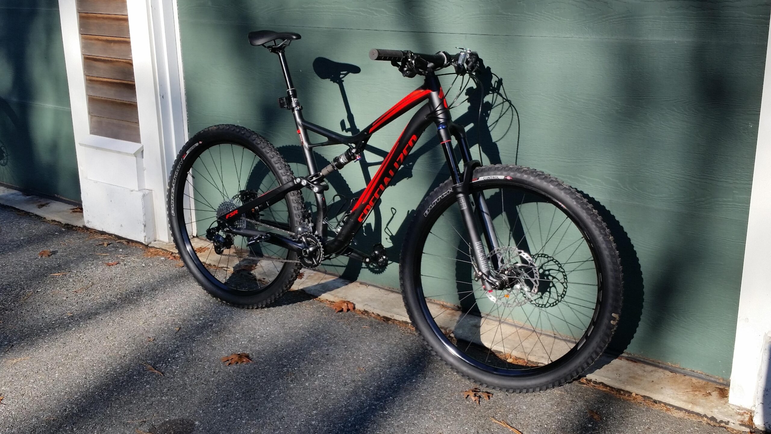 Specialized Stumpjumper FSR Comp 29er: A black and red mountain bike resting against a green wall, with a visible disc brake and wide tires. The bike is set on a paved surface, showcasing its modern design and features.