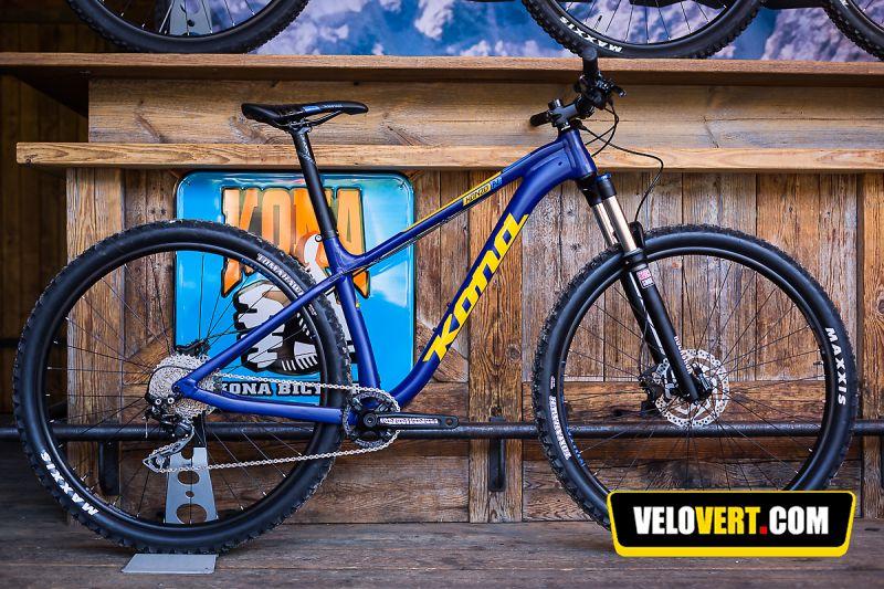 Kona Honzo: A blue Kona mountain bike displayed on a wooden shelf with a background featuring a colorful Kona sign. The bike has thick tires and a sturdy frame, suitable for off-road riding.