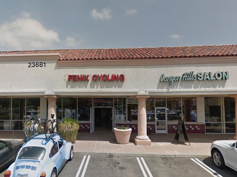 Front view of a shopping plaza featuring "Fenix Cycling" and "Canyon Hills Salon," with a light blue car parked in the foreground and bicycles on a roof rack. The building has a red tile roof and displays a welcoming sign.