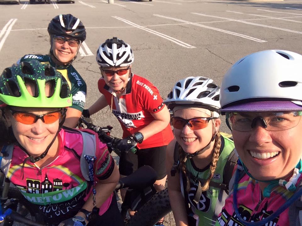 A group of five women wearing colorful cycling jerseys and helmets stand together, smiling for a selfie in a parking lot. They are equipped with biking gear, including sunglasses and gloves, and appear to be ready for a cycling adventure. The background shows empty parking spaces under a clear sky.