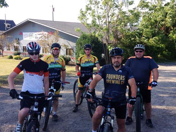 A group of five cyclists standing in a gravel area, wearing biking gear and helmets. They are posed on their mountain bikes, with a building and trees in the background. The cyclists are dressed in various colored jerseys, and some have logos on their clothing. The setting appears to be outdoors on a sunny day.