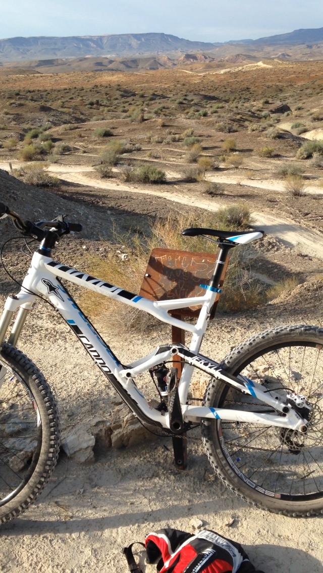 Cannondale Jekyll 3: A mountain bike with a white frame and blue accents rests on a dirt trail in a rugged, arid landscape. A small sign is partially obscured behind the bike, and the background features rolling hills and sparse vegetation under a clear sky. A black and red backpack lies on the ground nearby.