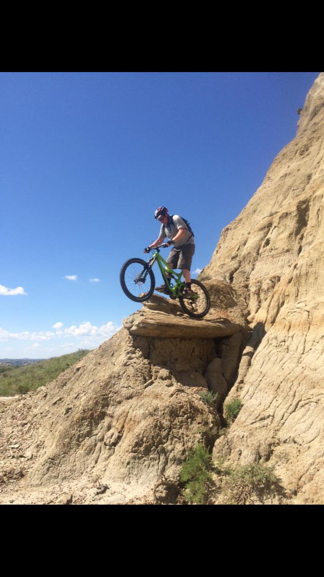Ibis Mojo HD3: A mountain biker navigating a rocky outcrop on a sunny day, with a clear blue sky in the background. The cyclist is poised on the edge of a dirt ledge, showcasing a green mountain bike and wearing a helmet and riding gear.