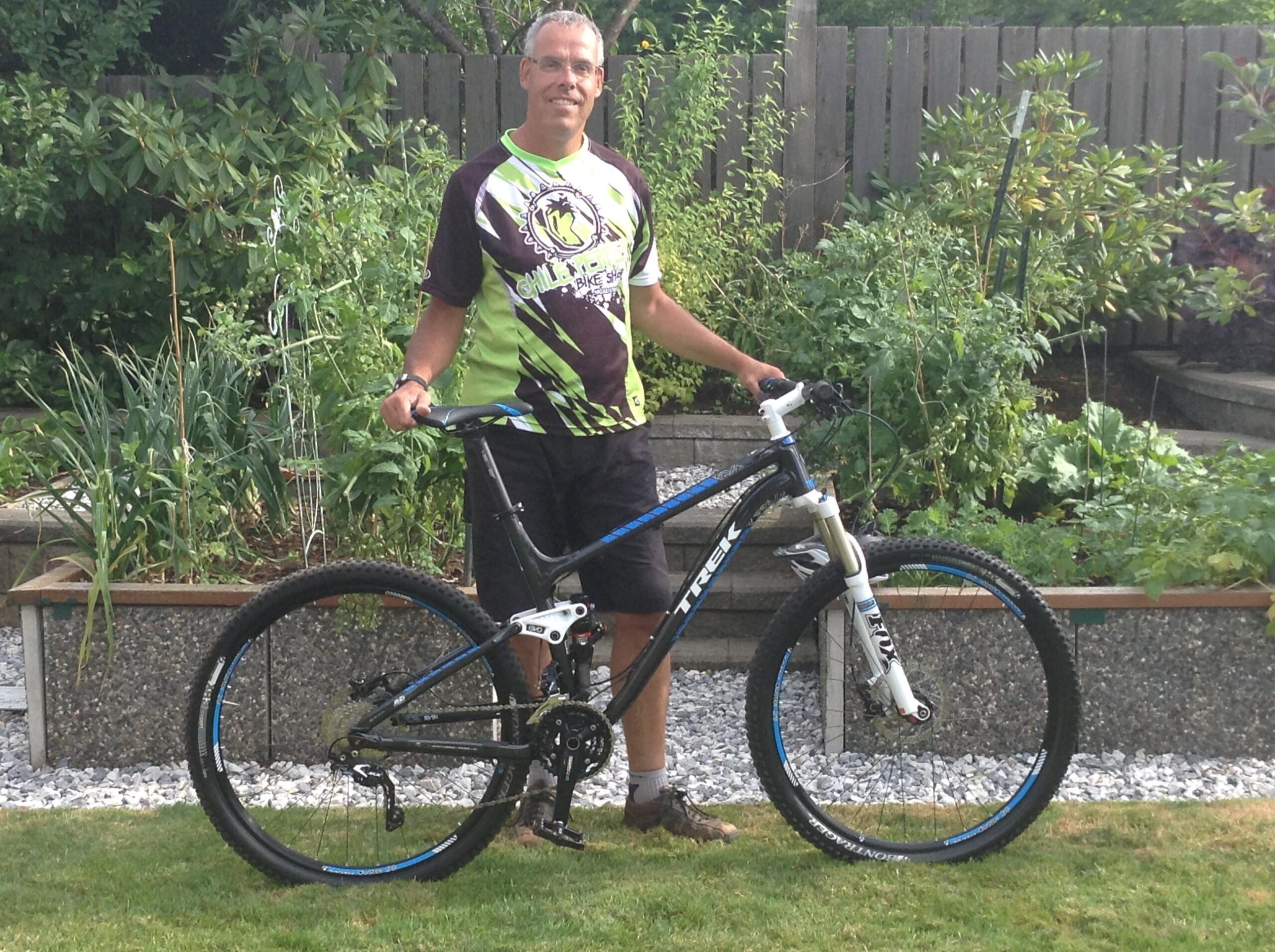 Trek Fuel EX 9 29: A person stands beside a mountain bike in a garden. The individual wears a green and black bike jersey and shorts, smiling at the camera. The background features various plants and a wooden fence, with gravel and greenery surrounding the bicycle. The bike is black with blue accents.