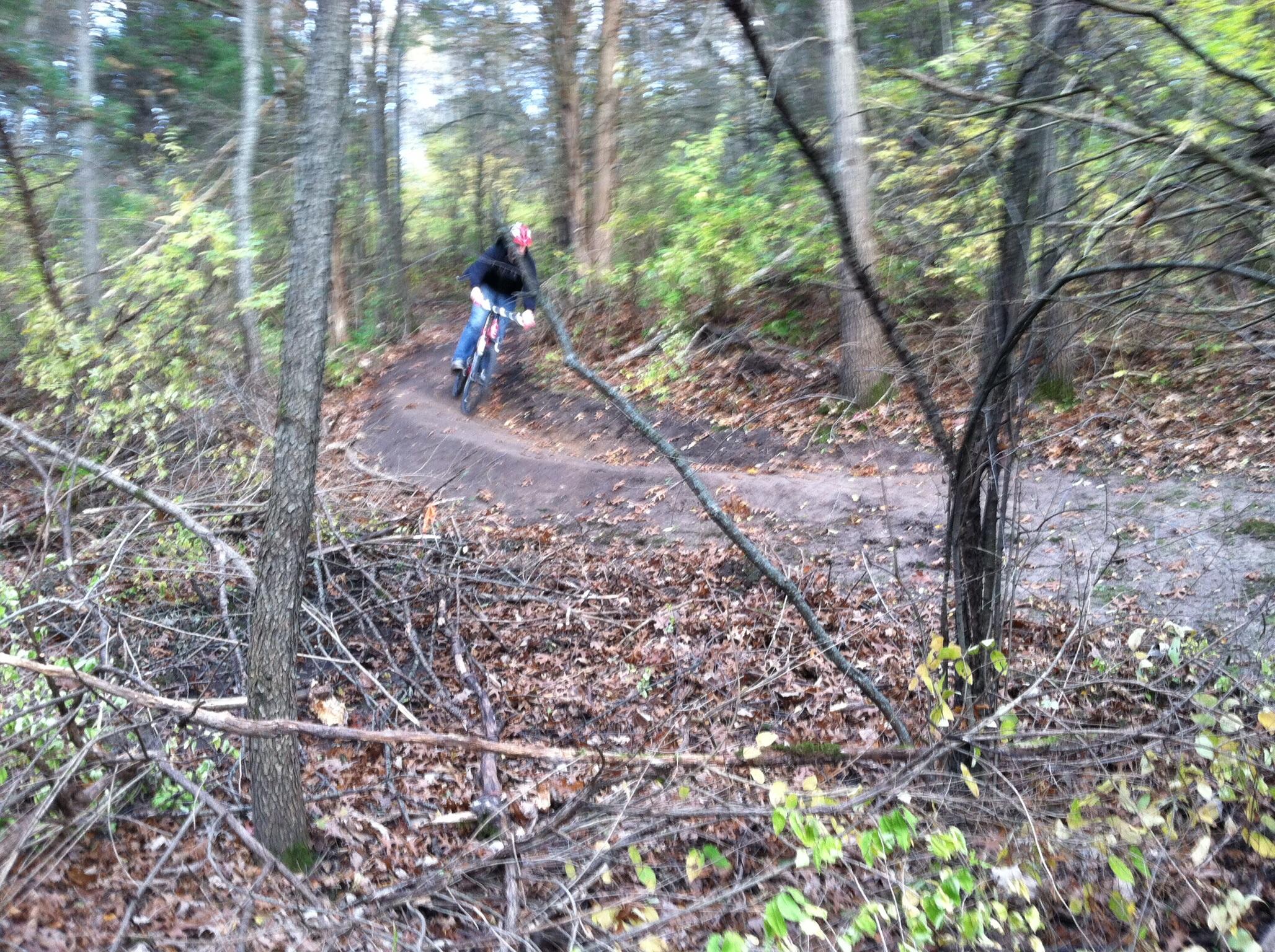 A mountain biker riding along a winding dirt trail through a forested area, surrounded by trees and fallen branches, with autumn foliage on the ground. The image is slightly blurred, suggesting movement. Silver Lake Park mountain bike trail.