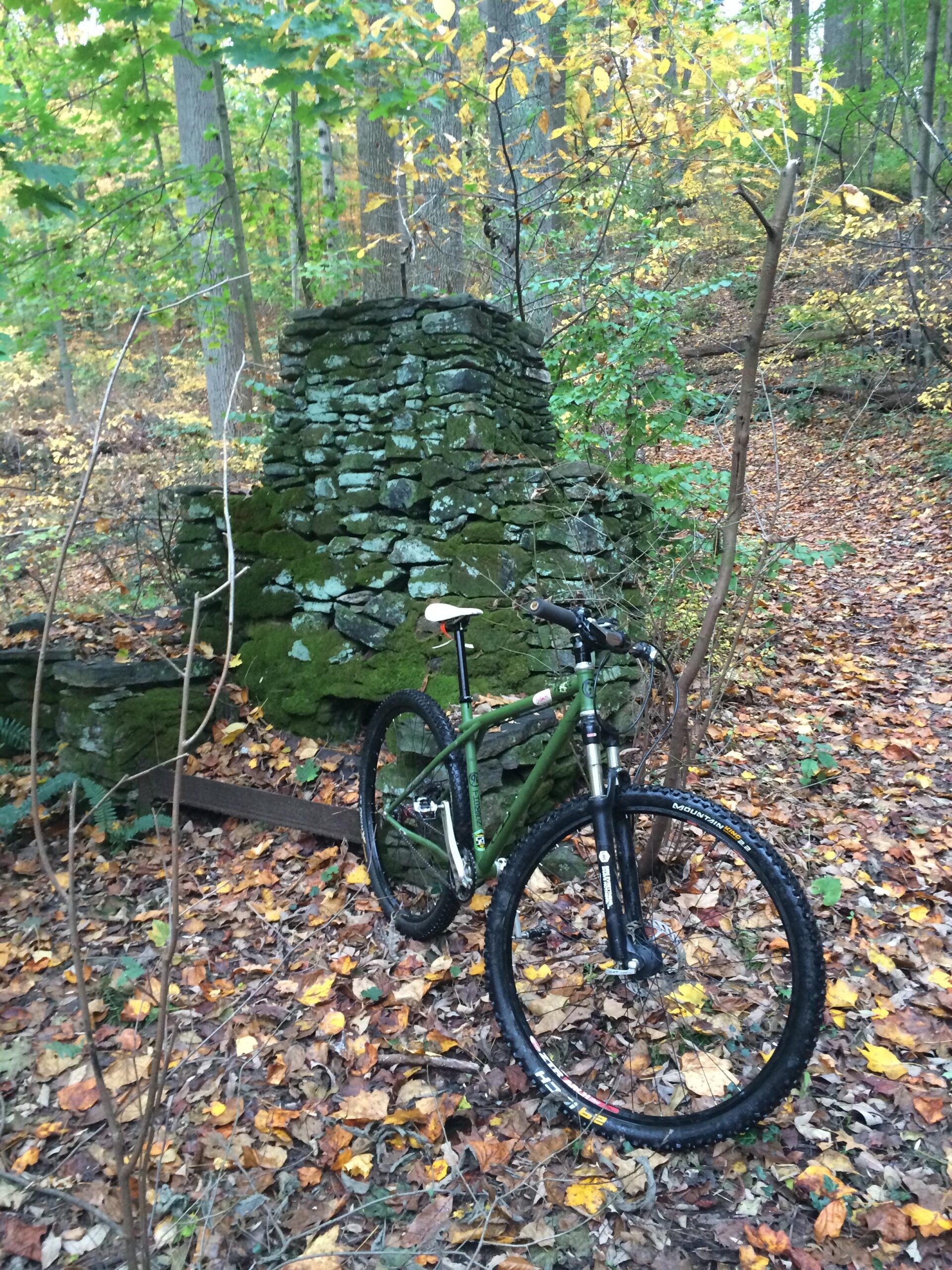A mountain bike parked beside a weathered stone structure in a wooded area, surrounded by autumn leaves and trees with yellow and green foliage. Brandywine State Park mountain bike trail.