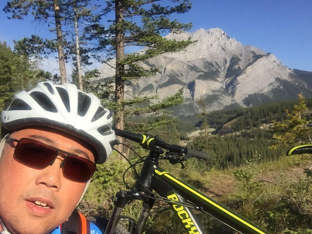 A person wearing a white bicycle helmet and sunglasses is taking a selfie in a scenic outdoor setting, with a mountain range and dense forest in the background. A mountain bike is visible beside them. Tunnel Mountain Trail System mountain bike trail.