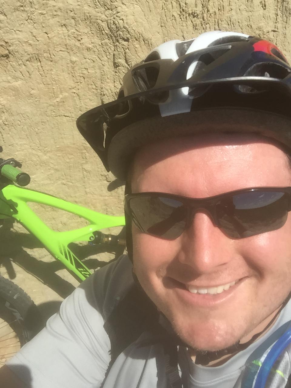 Ibis Mojo HD3: A person wearing a black and white bicycle helmet and sunglasses smiles for a selfie, with a bright green mountain bike partially visible in the foreground. The background features a textured, sandy surface, suggesting an outdoor setting.