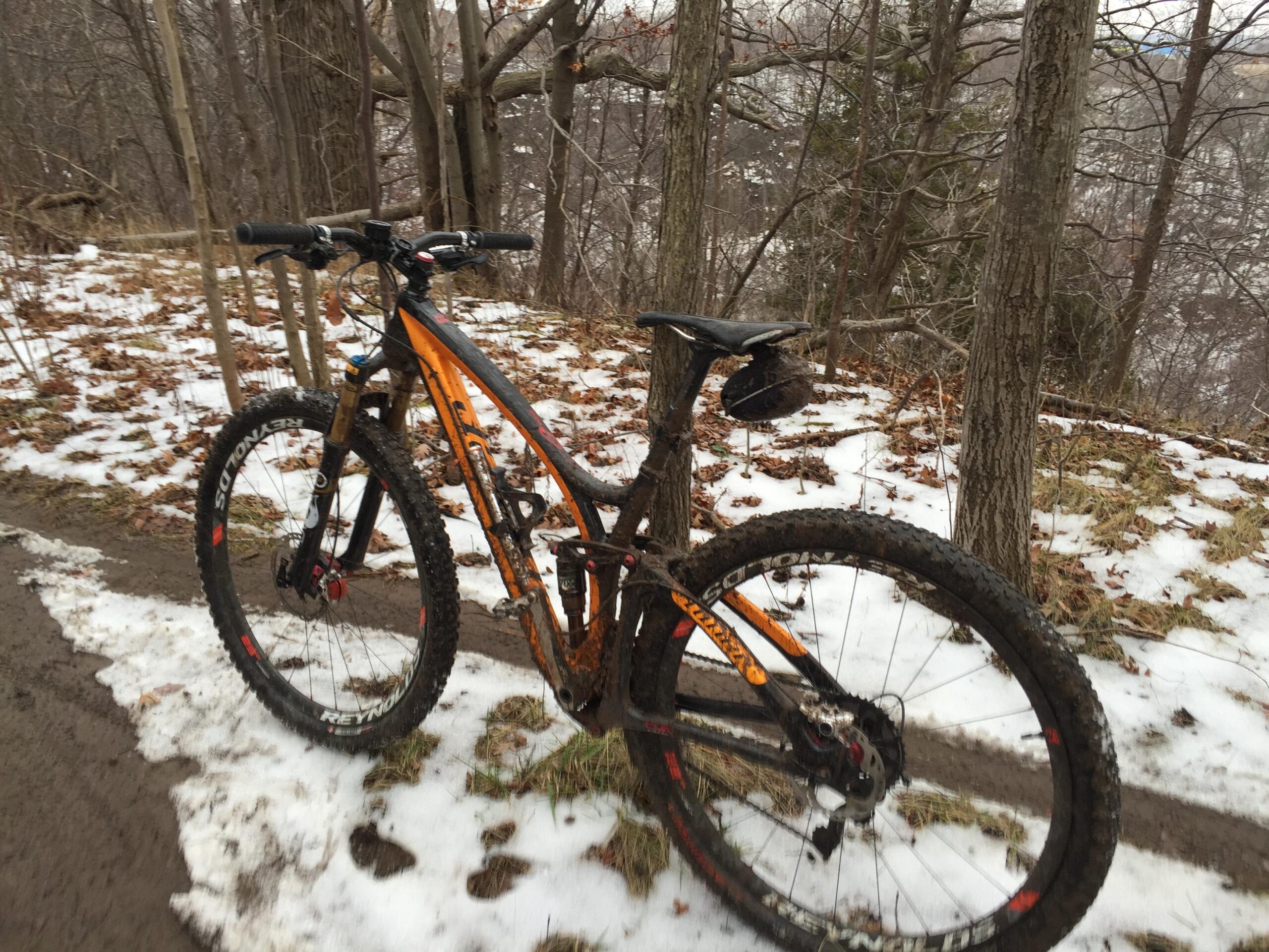 Niner Jet 9 RDO: Mountain bike parked on a trail surrounded by trees and partially covered in snow. The bike is orange and black, with mud splattered on the frame and tires, indicating recent use. The background features fallen leaves and a glimpse of a snowy landscape.