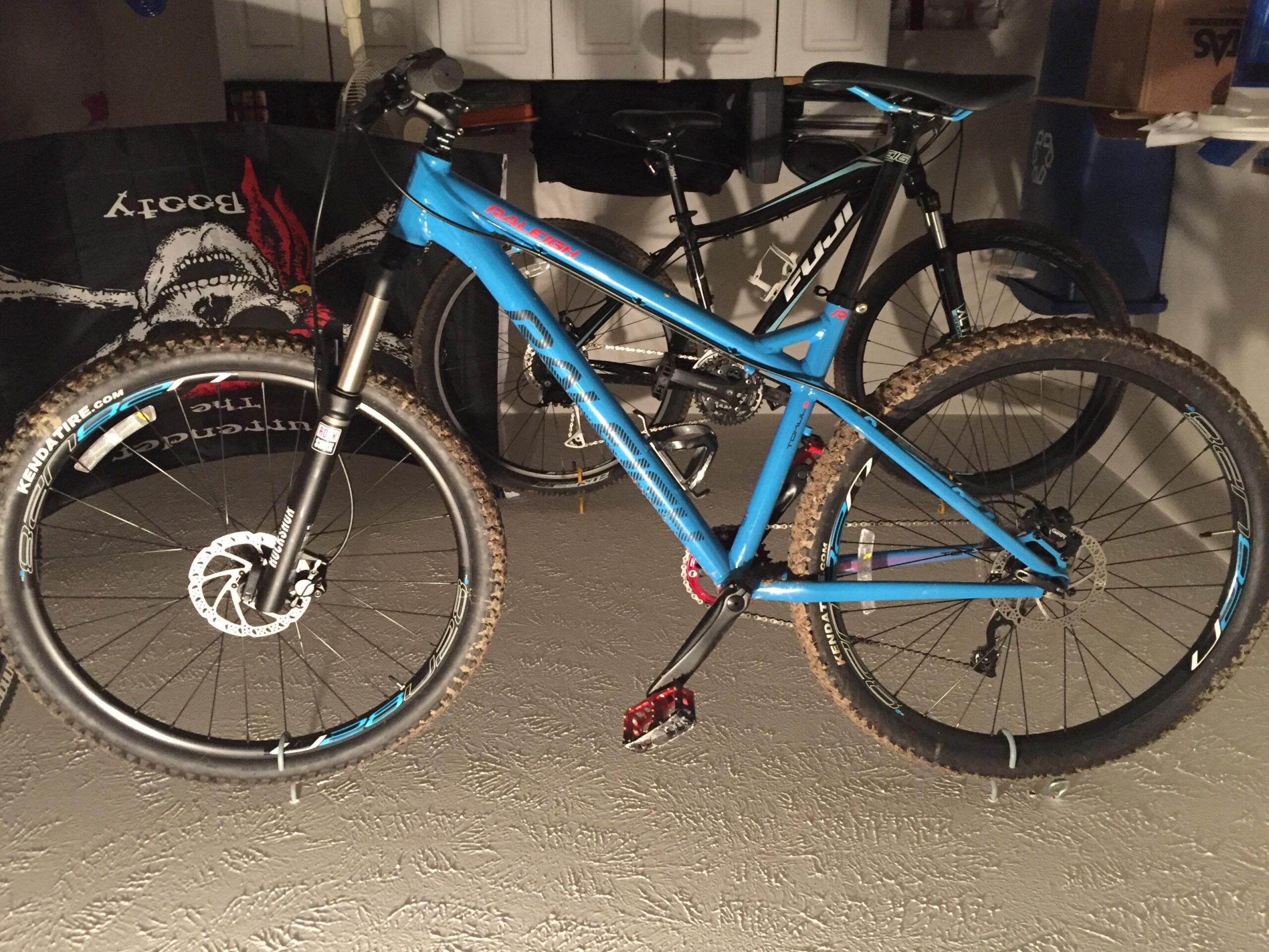 Raleigh Tokul: A blue mountain bike with a front suspension fork is positioned next to a black mountain bike in a garage setting. The foreground bike features knobby tires with visible dirt, a disc brake system, and a sleek frame design. In the background, the black bike is partially obscured. The garage walls contain various items, including a dark banner and storage boxes.