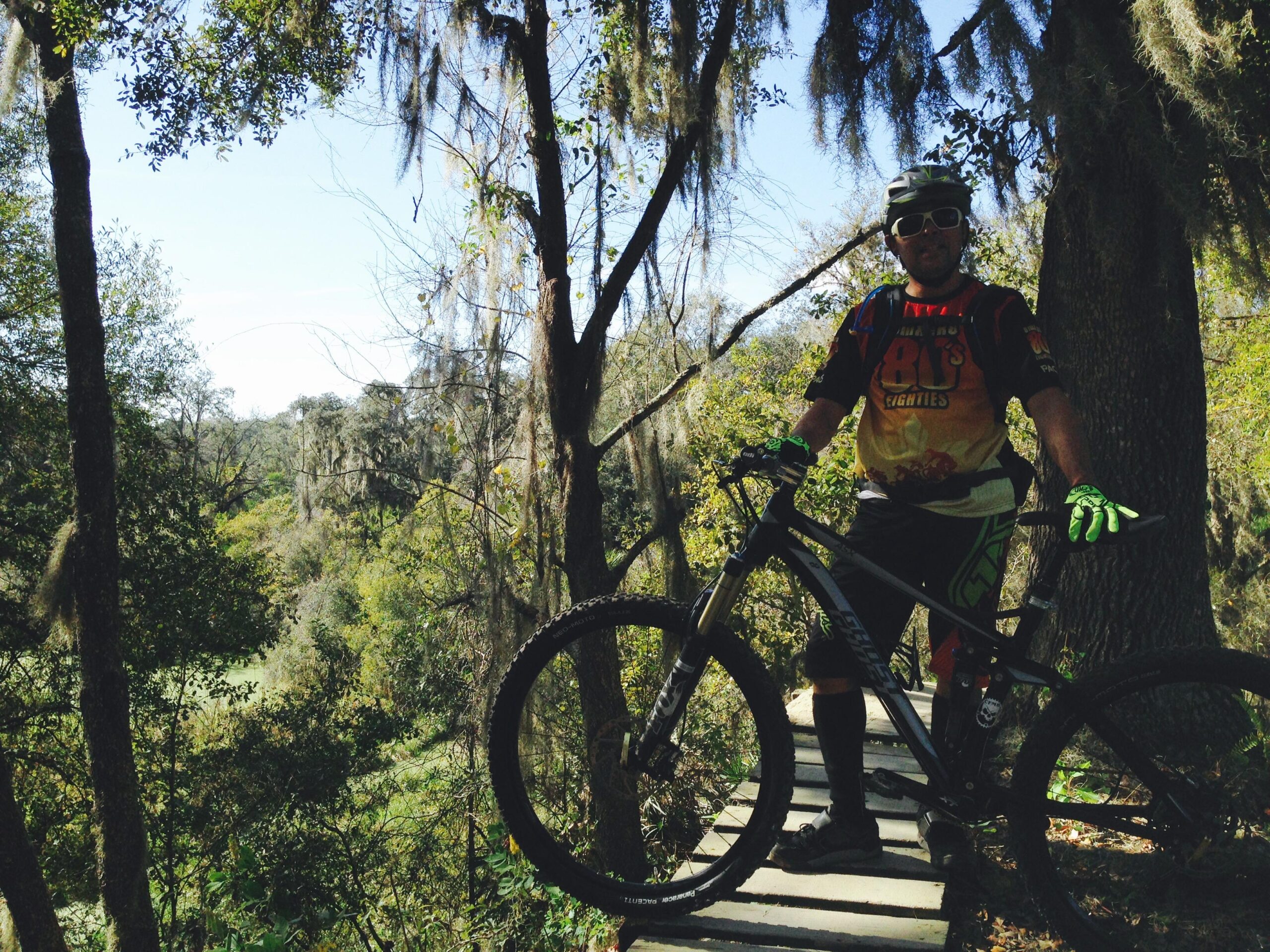 Ghost Ghost ASX 7500: A mountain biker stands on a wooden bridge surrounded by lush green trees and foliage. He wears a colorful cycling jersey, gloves, and a helmet, holding onto his bike as he enjoys the outdoor scenery. The bright blue sky is visible through the leaves above, indicating a sunny day.