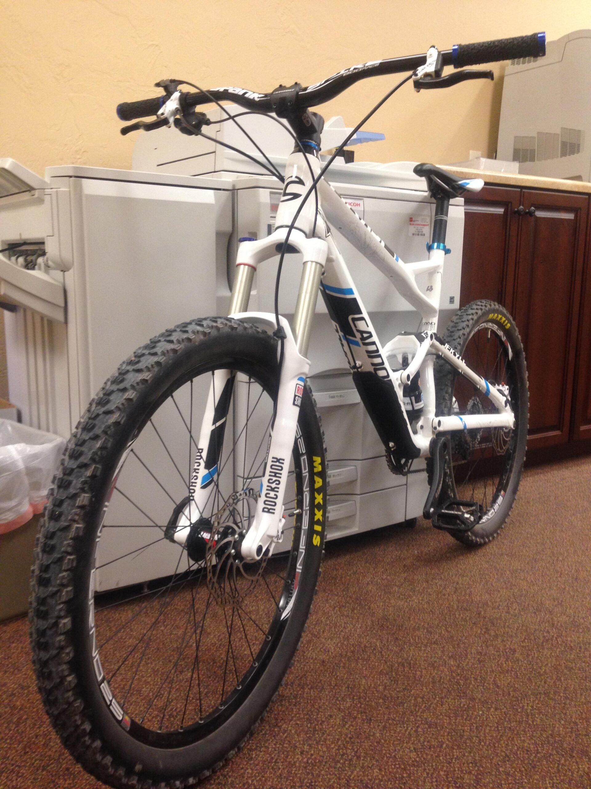 Cannondale Jekyll 3: A white mountain bike with black and blue accents is leaning against a copier in an office setting. The bike features large tires and a sturdy frame, with the handlebars positioned at an angle. In the background, there are office furniture and equipment visible, including a wood cabinet and another copier.
