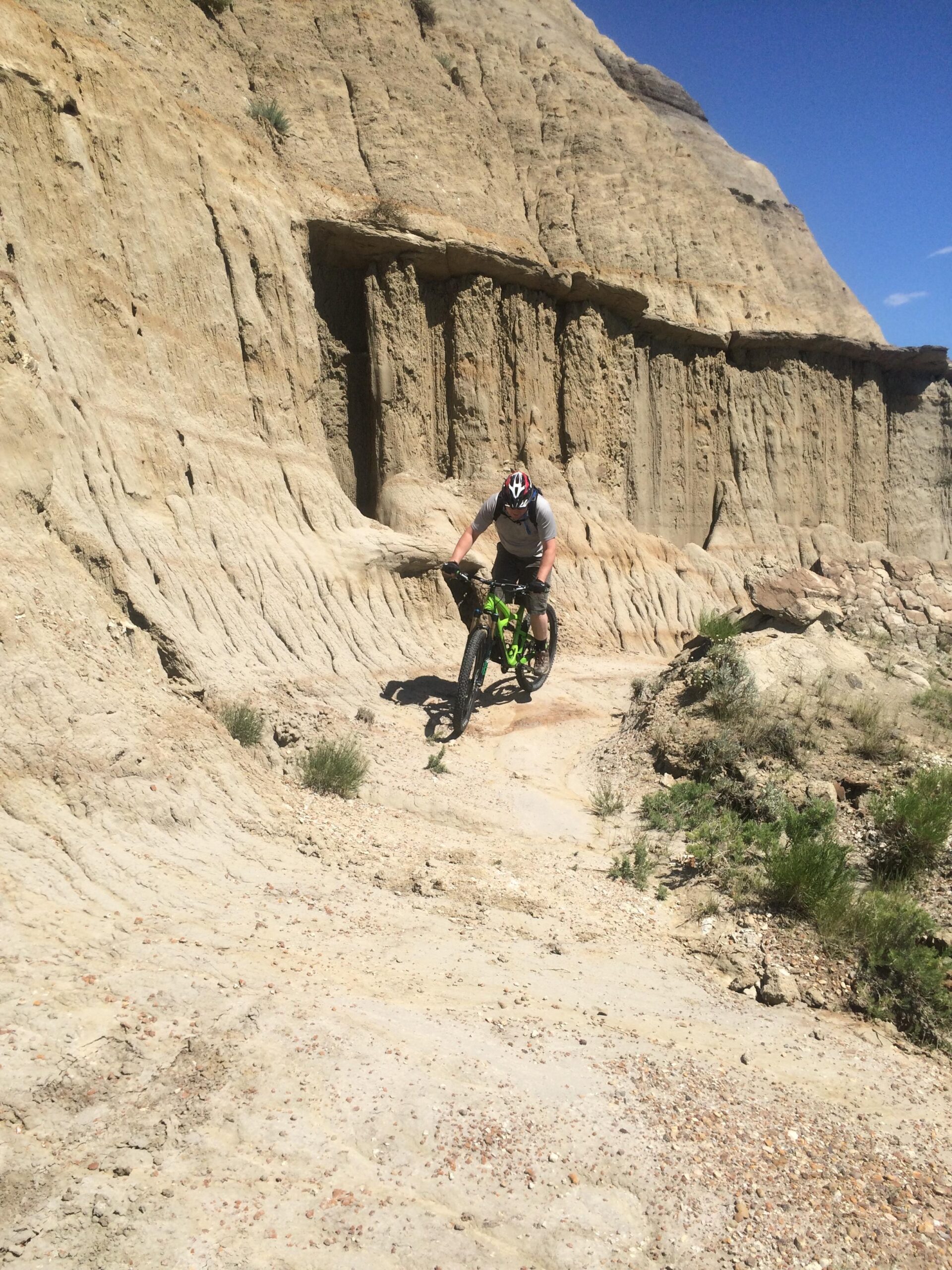 Ibis Mojo HD3: A mountain biker navigates a rugged, dry terrain with steep, rocky cliffs in the background under a clear blue sky. The cyclist is wearing a helmet and riding a green bike, emphasizing the challenging outdoor environment.