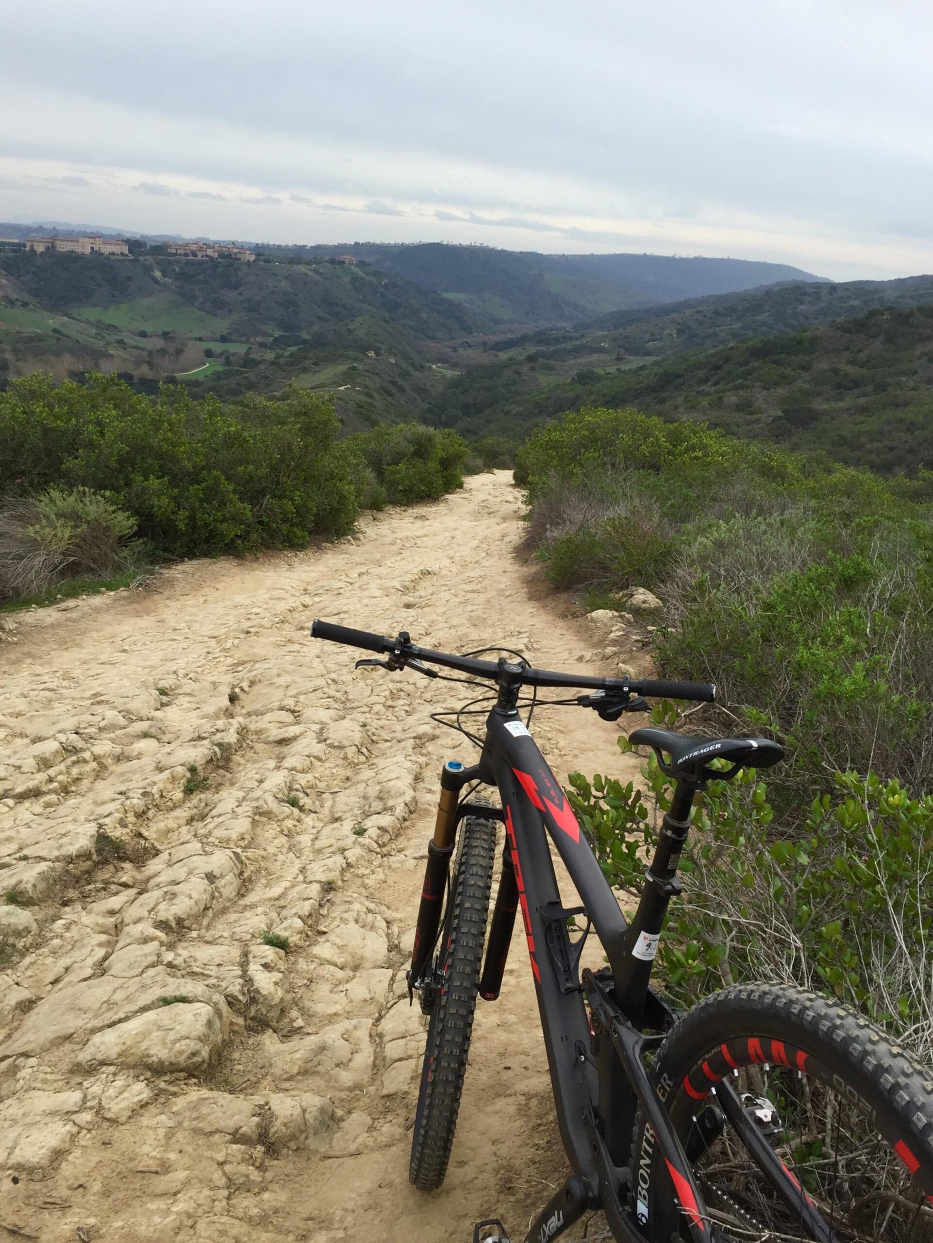 A mountain bike parked on a rocky dirt trail overlooking a lush green valley, with hills and a cloudy sky in the background. Aliso and Wood Canyons Wilderness Park mountain bike trail.