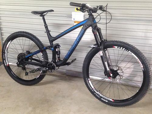 Transition Scout: Alt text: A black and blue mountain bike with thick tires is leaning against a wall. The bike features a full suspension system, a sturdy frame, and has various components visible, including the handlebars and gears.