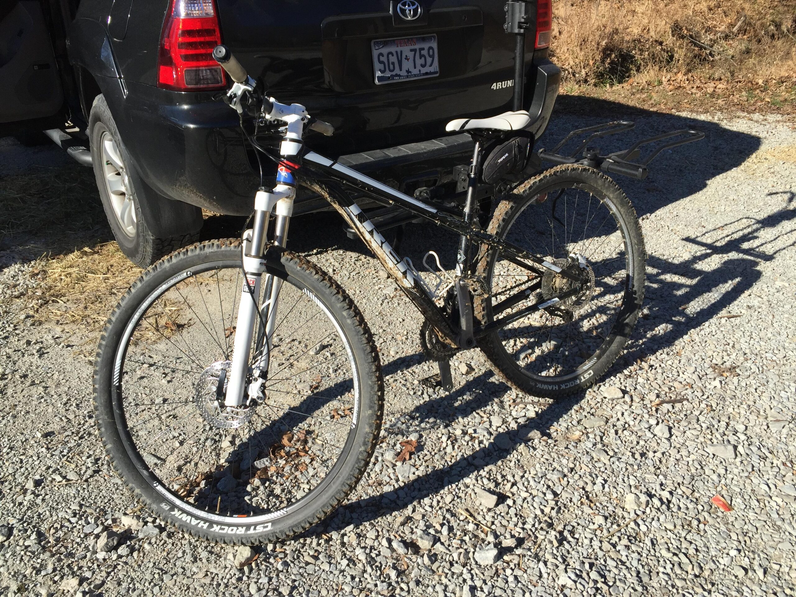 Trek X-Caliber 8: A black mountain bike with white front forks is parked on gravel next to a dark SUV. The bike features knobby tires, a rear rack, and a dirt-speckled finish, indicating recent use. The background shows dry grass and foliage.