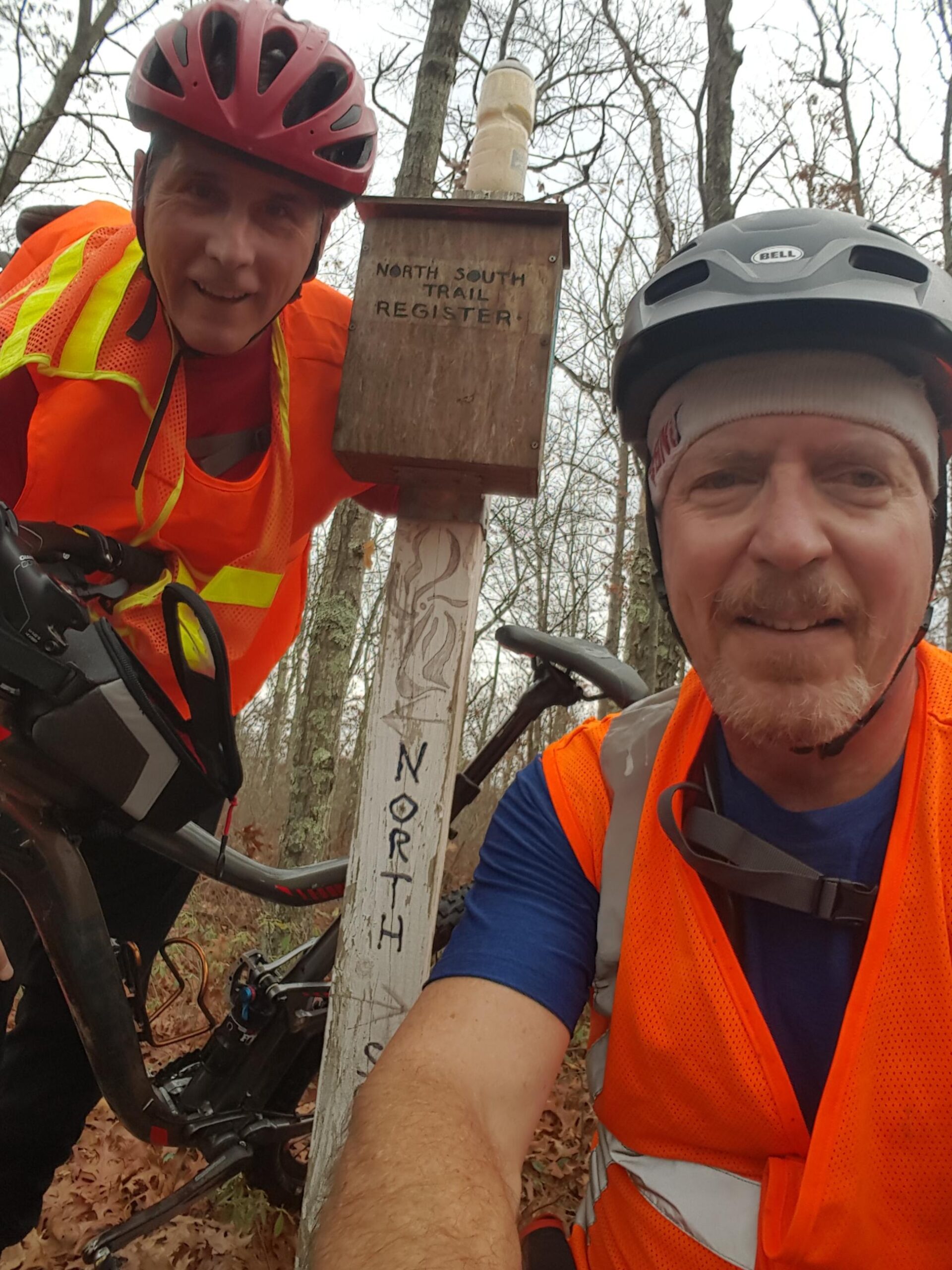 Two cyclists wearing bright orange vests and helmets pose for a selfie at a trail intersection marked "North South Trail Register." The background features a wooded area with trees and fallen leaves. One cyclist is leaning on a mountain bike, while both are smiling at the camera. North South Trail mountain bike trail.